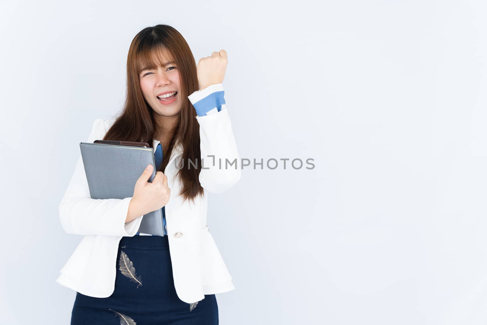 Smiling Asian business woman wearing a medical face mask holding the notebook and showing fist fighting over grey background. Back to the normal concept.