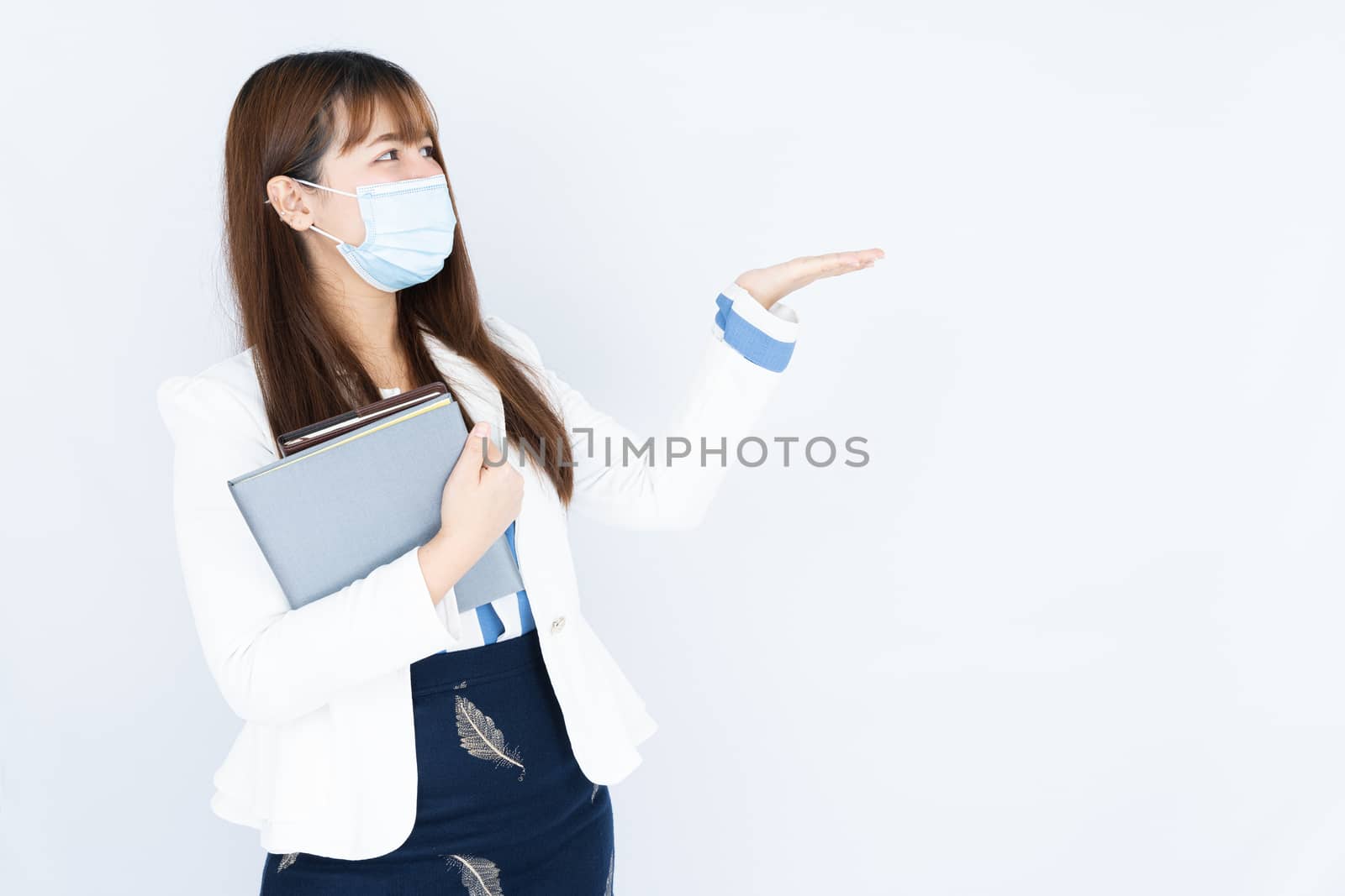 Smiling Asian business woman wearing a medical face mask holding the notebook and pointing hand to side blank space over grey background. Back to the normal concept.