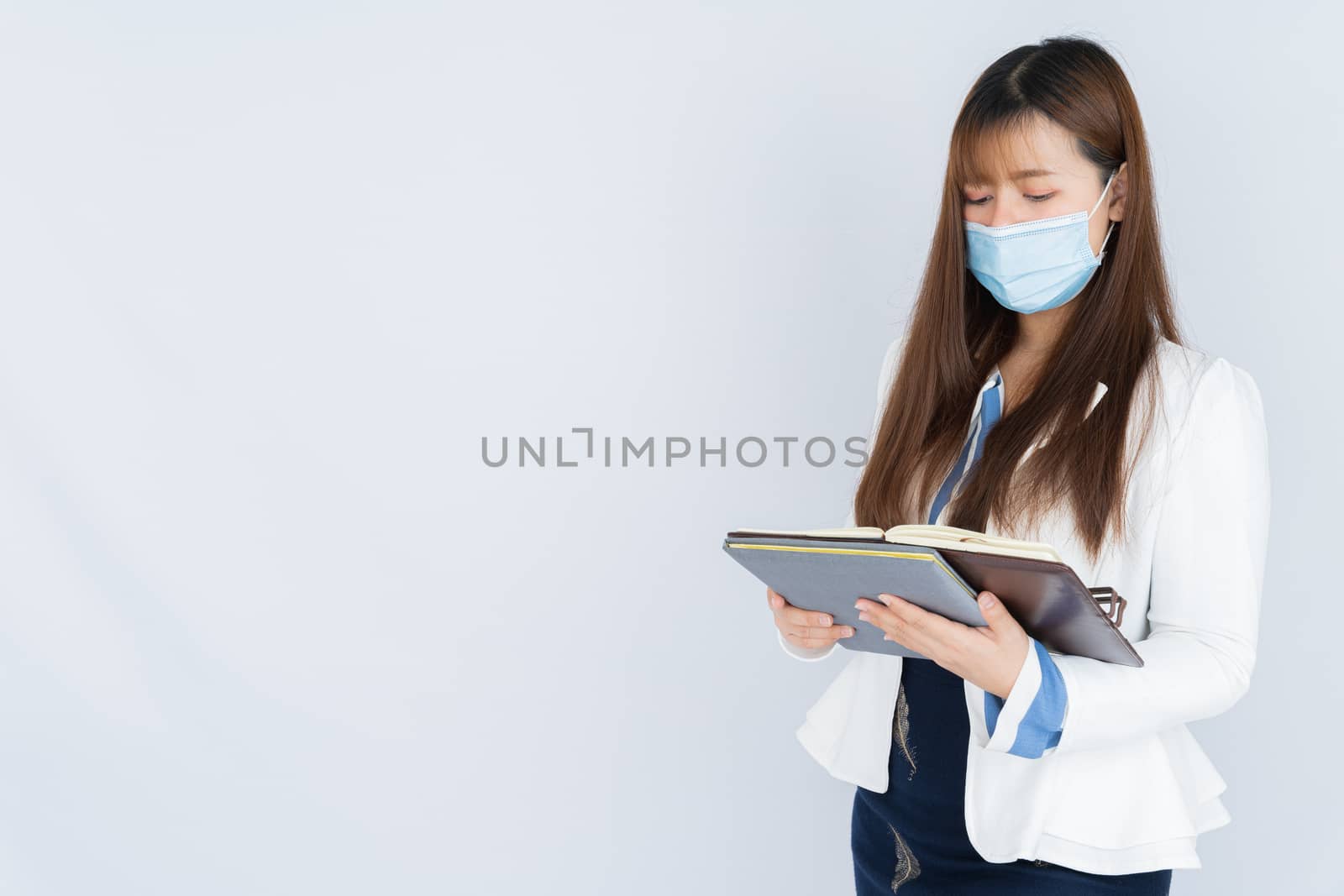 Asian business woman wearing a medical face mask and looking at the notebook that she holding over grey background. Back to the normal concept.