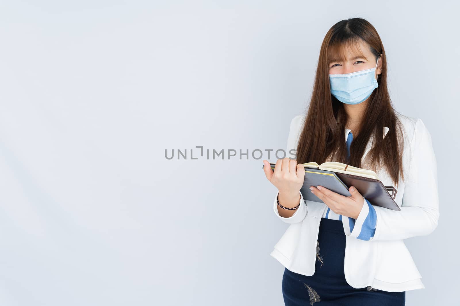 Smiling Asian business woman wearing a medical face mask holding the notebook and looking at the camera over grey background. Back to the normal concept. by mikesaran