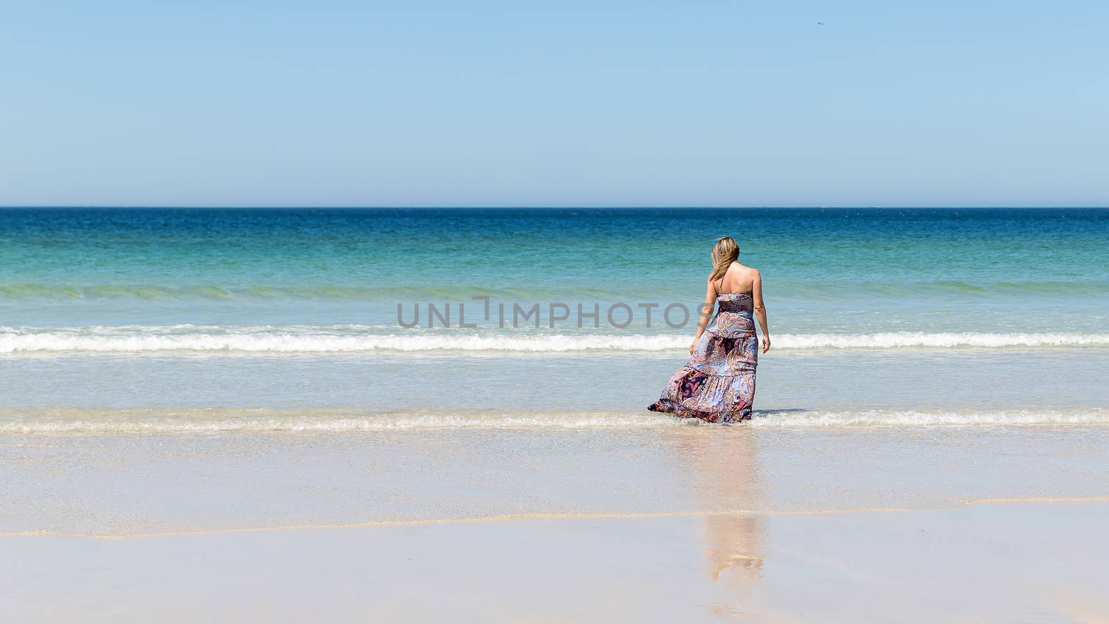 Middle-aged blonde woman in a long dress relaxes on the beach facing the sea
