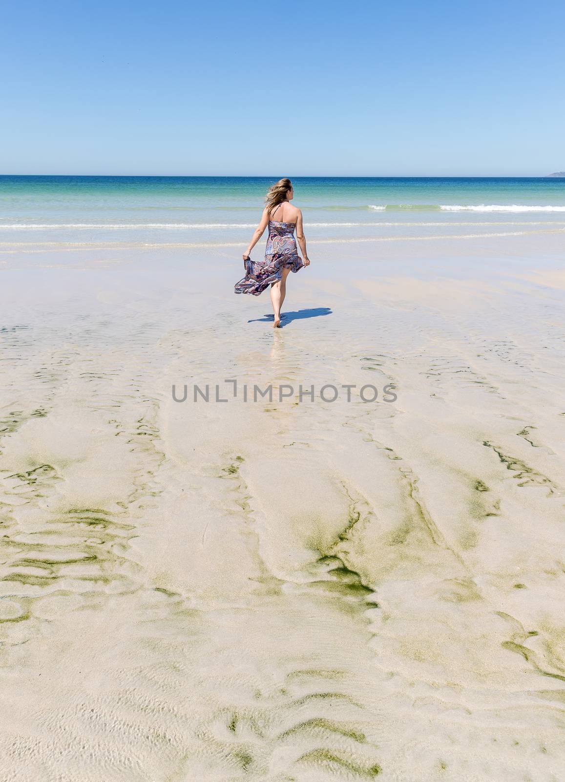 Middle-aged blonde woman in a long dress walking on the beach towards the sea