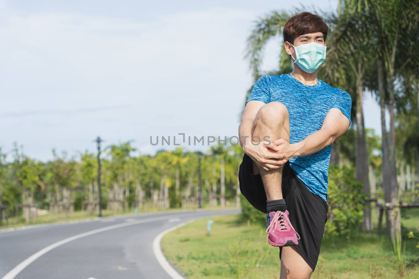 Young male wearing medical mask and stretching his legs before workout training session at the park. Stay in fit during quarantine in the days of the Corona Virus or Covid-19