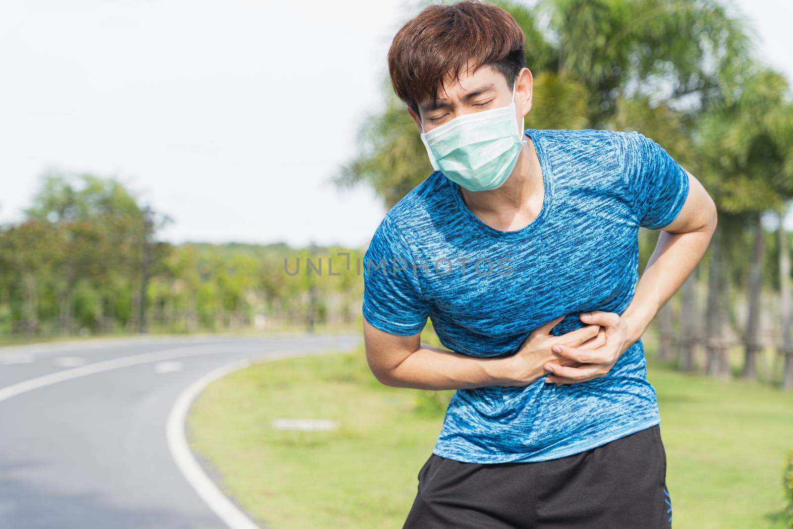 Young male wearing medical mask have stomach ache while workout at the park. Stay in fit during quarantine in the days of the Corona Virus or Covid-19