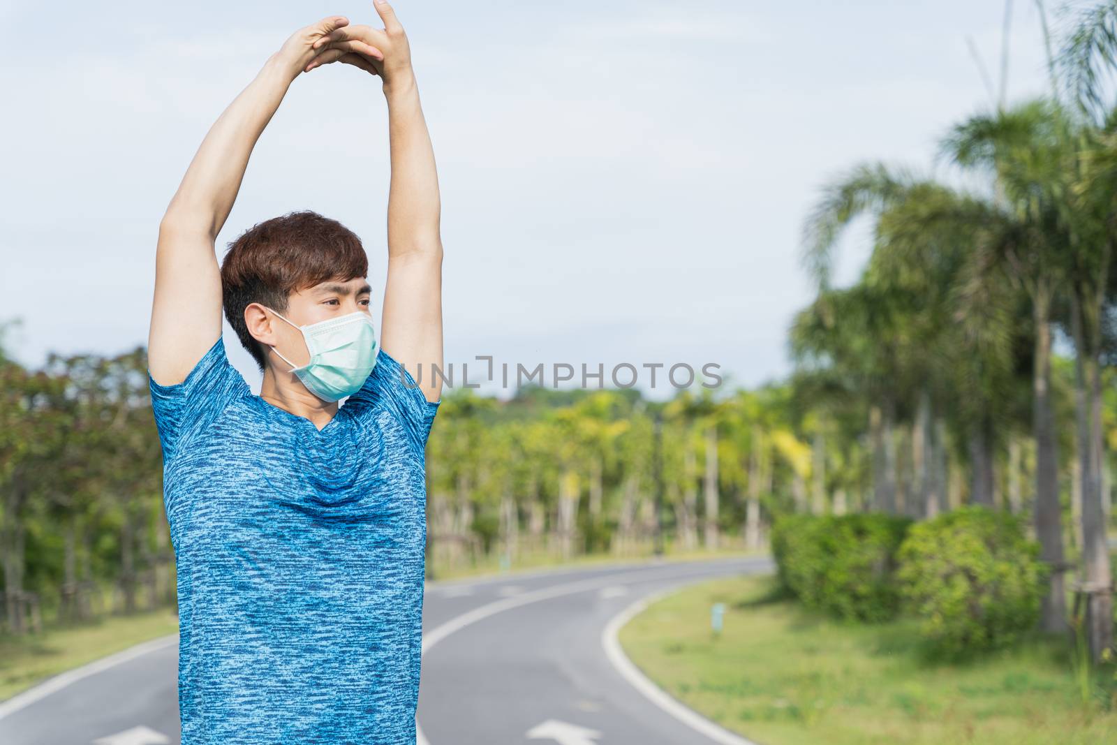 Young male wearing medical mask and stretching his arms before workout training session at the park. Stay in fit during quarantine in the days of the Corona Virus or Covid-19