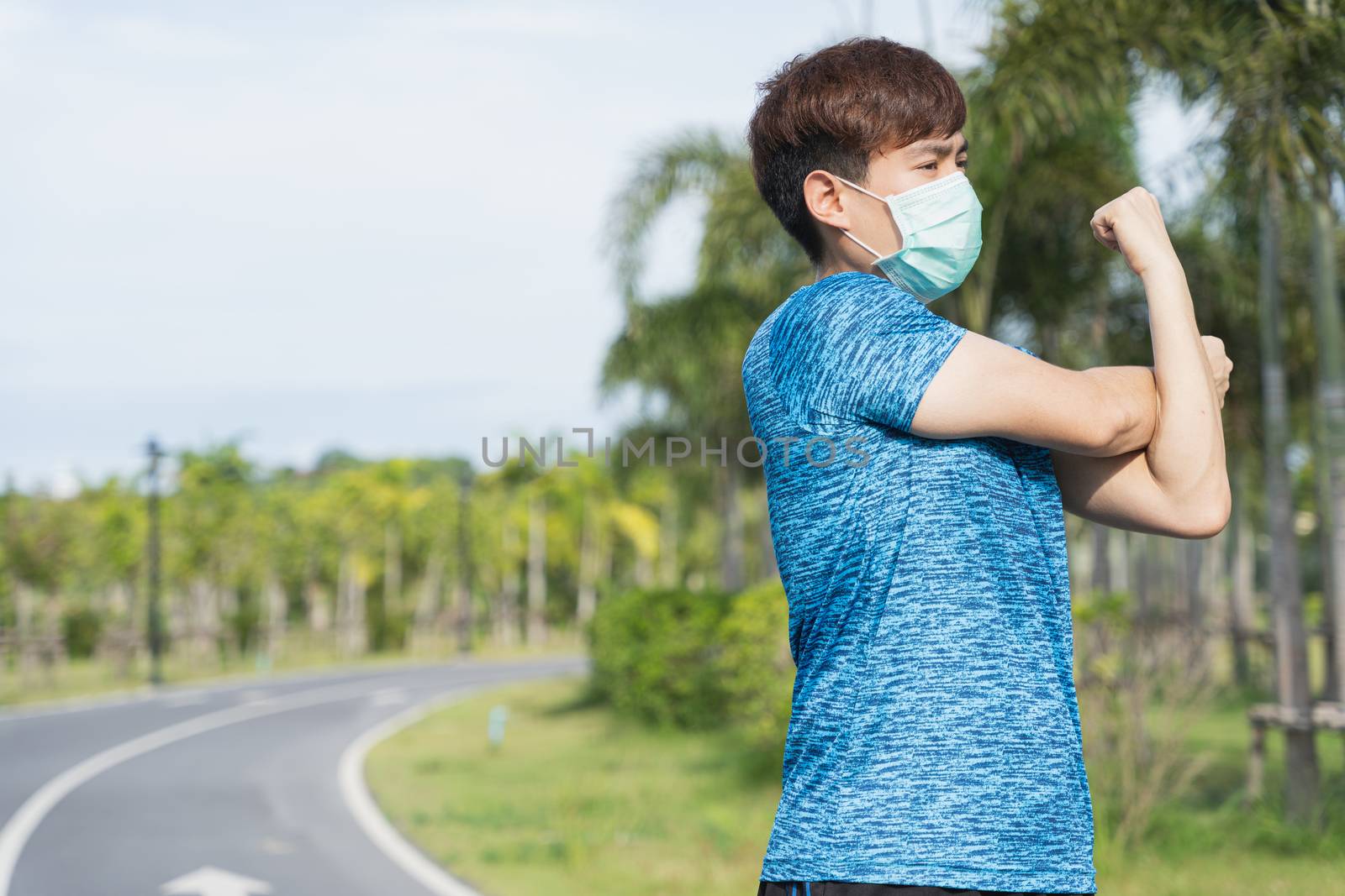 Young male wearing medical mask and stretching his arms before workout training session at the park. Stay in fit during quarantine in the days of the Corona Virus or Covid-19