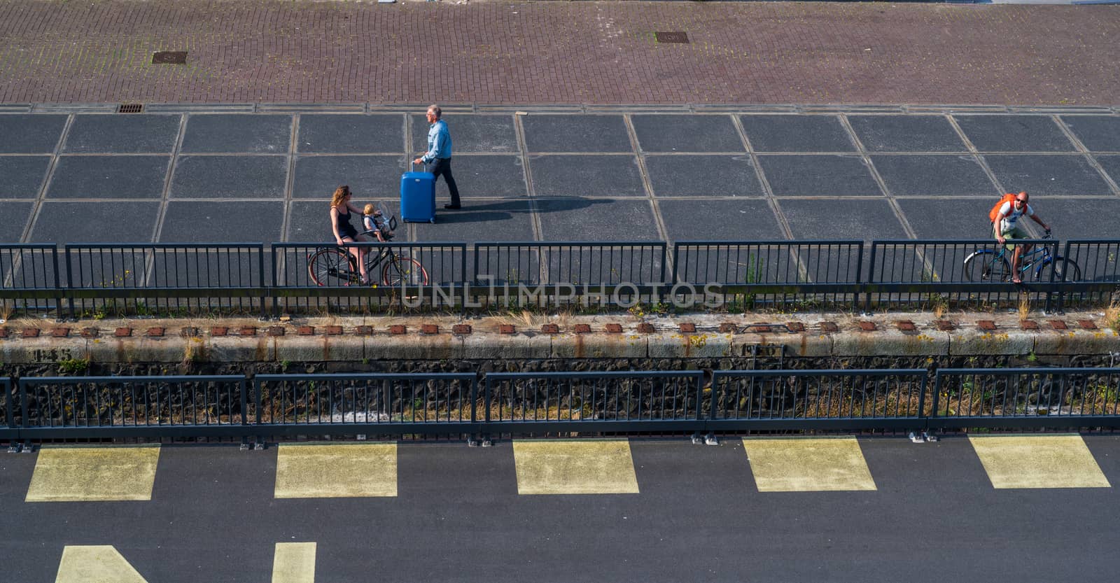 Amsterdam, the Netherlands — July 28, 2019. A man walks by the pier with a blue suitcase; bicycle riders head in the opposite direction.