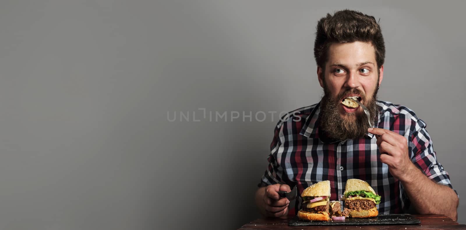 Young man eating fresh tasty large hamburger, gray background with copy space