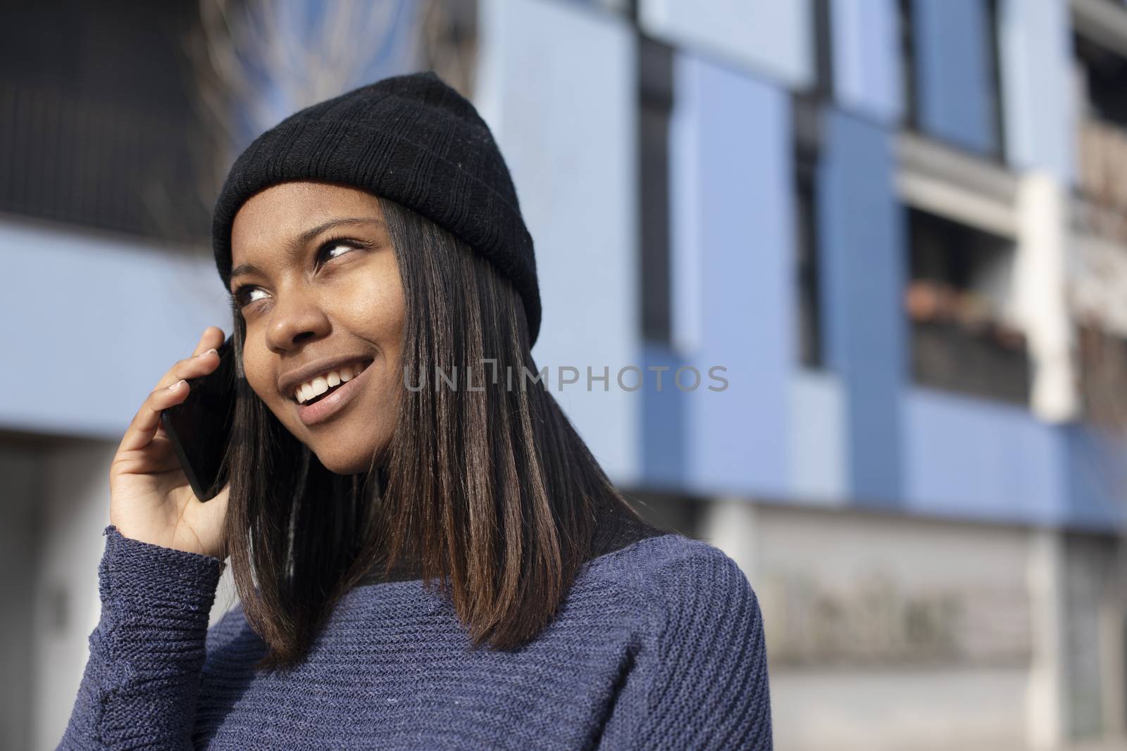 Portrait of African American young beautiful woman wearing a wool cap standing on the street while using a mobile phone by raferto1973