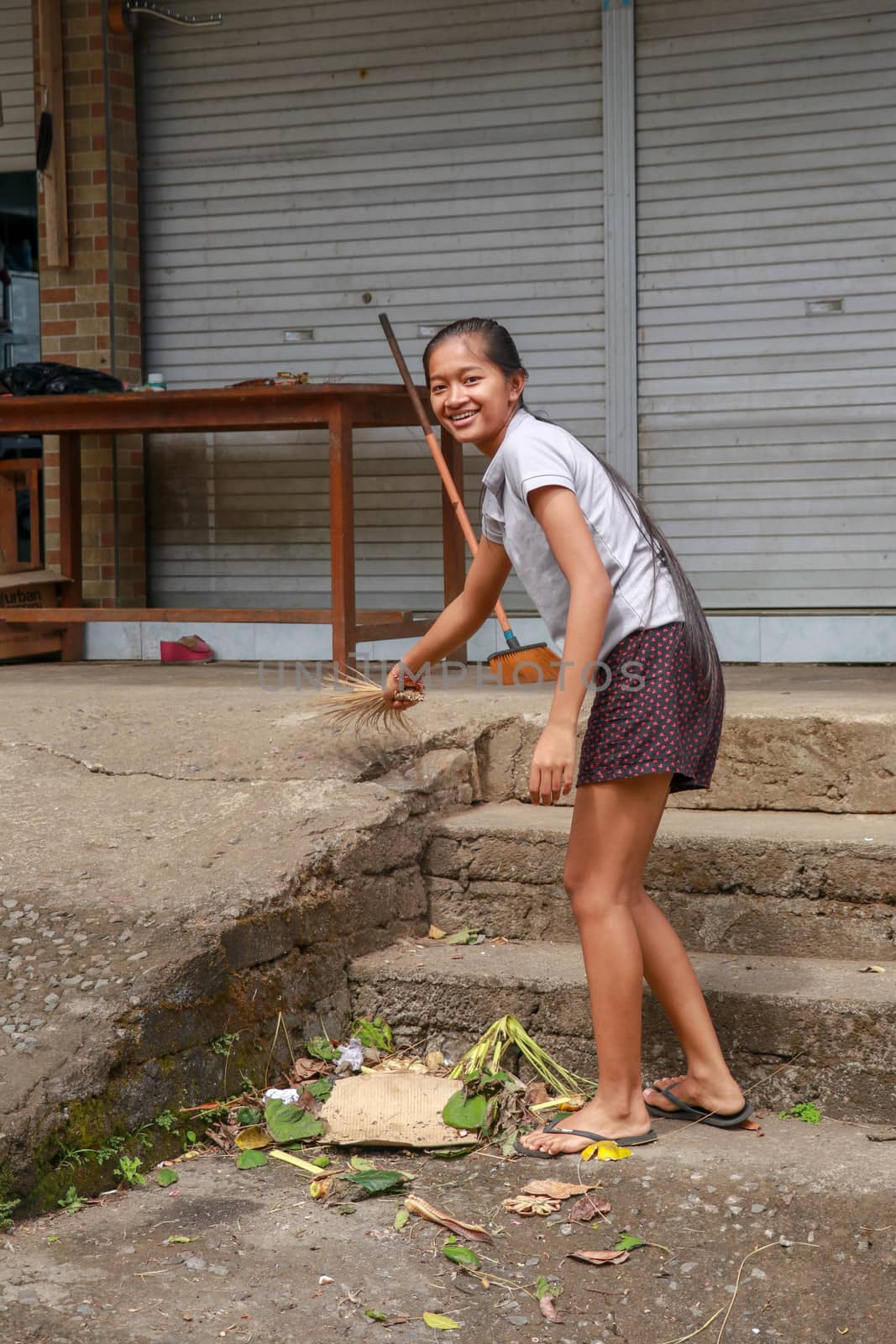 Woman cleaning service with broom stick and dustpan clean up the road. Balinese girl sweeps garbage and dry leaves in front of the house. Bali, Indonesia by Sanatana2008