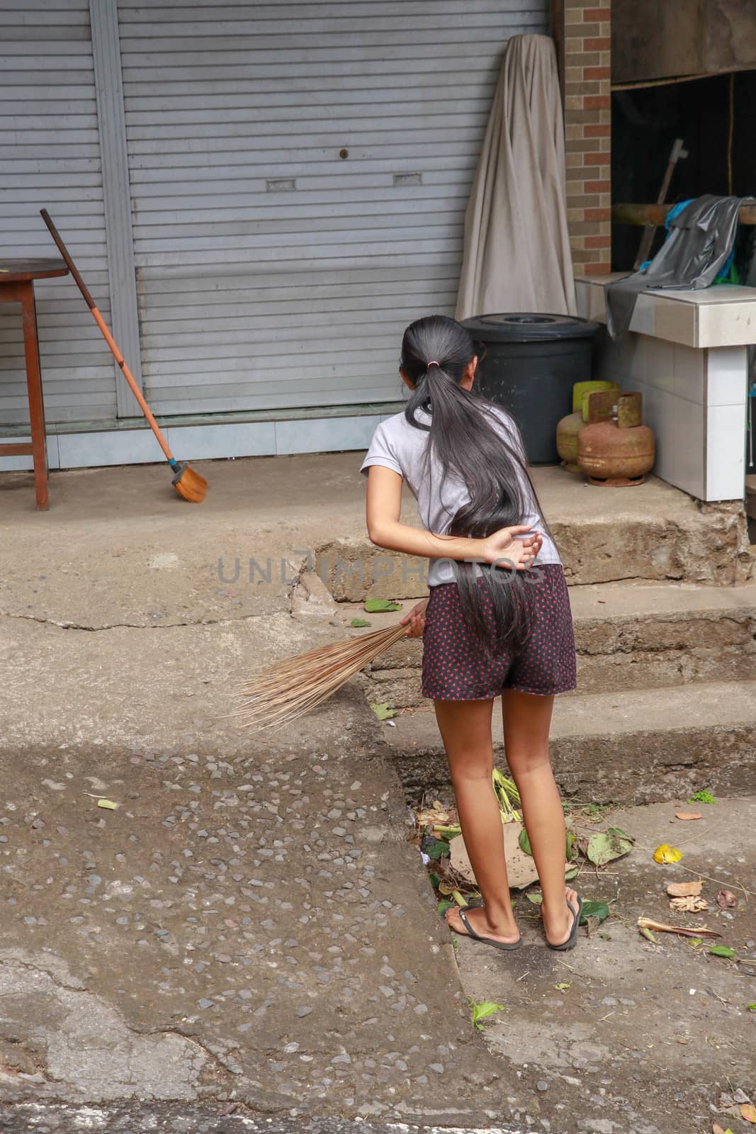 Woman cleaning service with broom stick and dustpan clean up the road. Balinese girl sweeps garbage and dry leaves in front of the house. Bali, Indonesia by Sanatana2008
