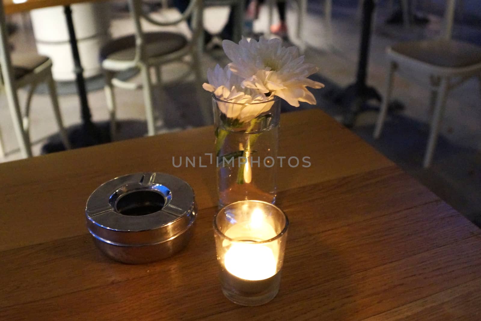 lighted candle, chrysanthemum flower and ashtray on a wooden table of a street cafe in the evening by Annado