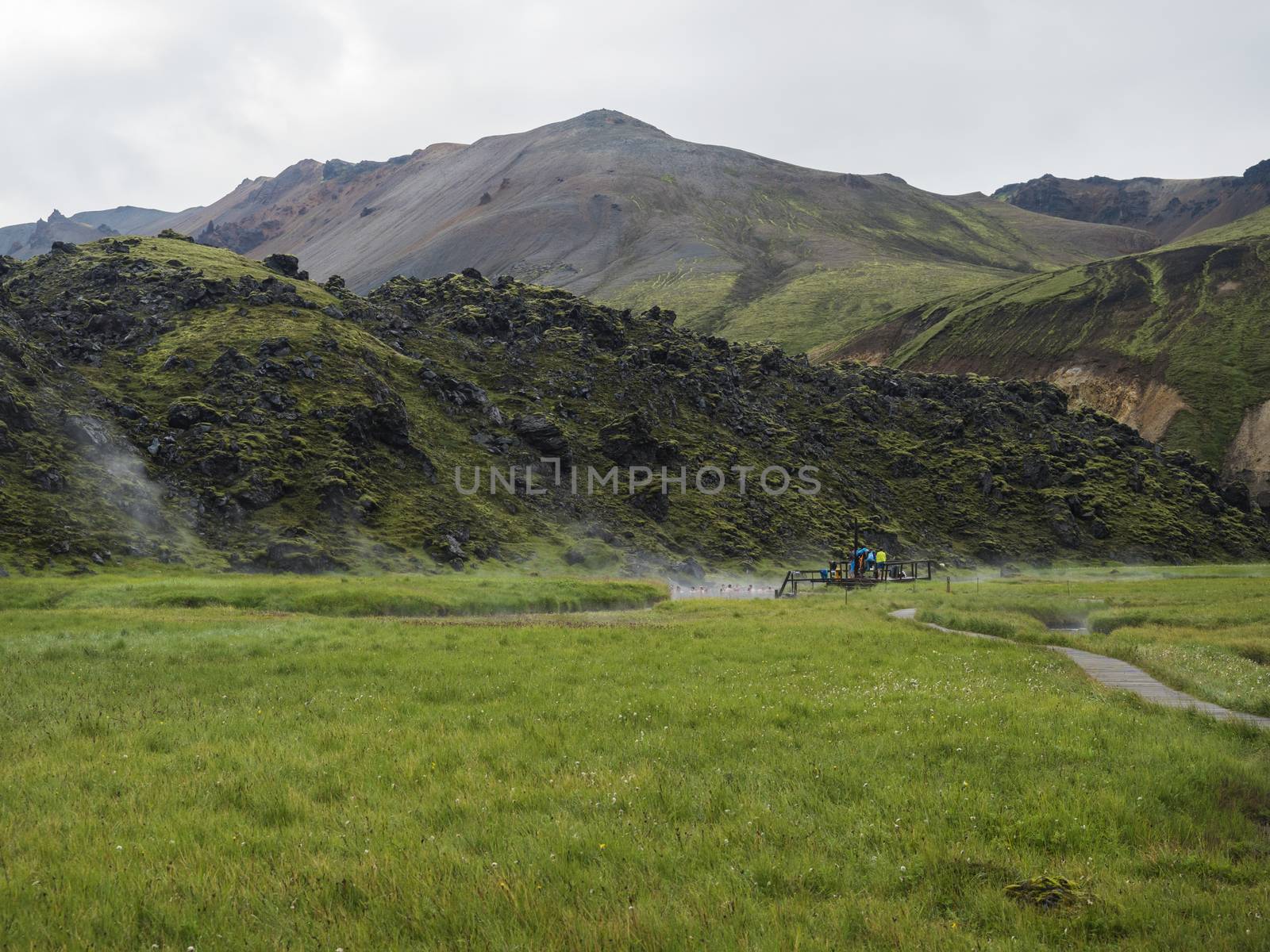 Group of tourist people relaxing in a natural hot spring in thermal baths in Landmannalaugar camp site, Iceland. Grass meadow, lava fields and mountains in background.
