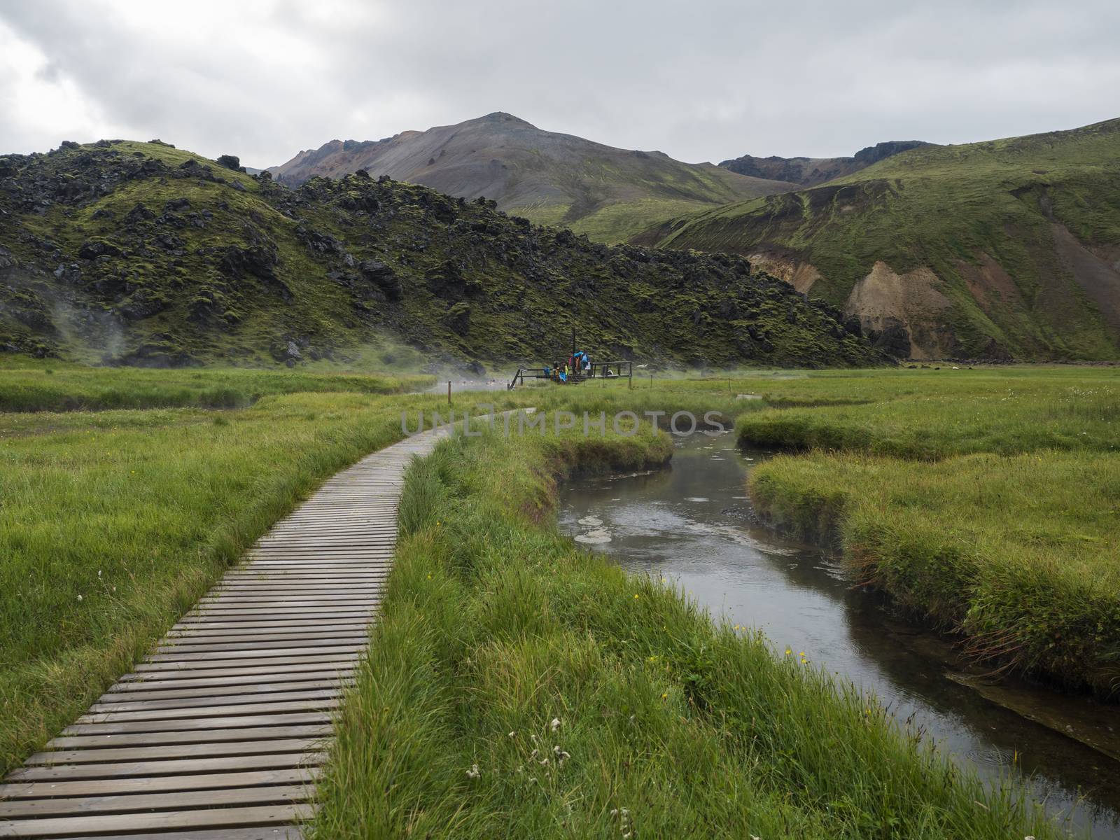 Wooden path to natural hot spring with group of tourist people relaxing in a thermal baths in Landmannalaugar camp site, Iceland. Grass meadow, lava fields and mountains in background.