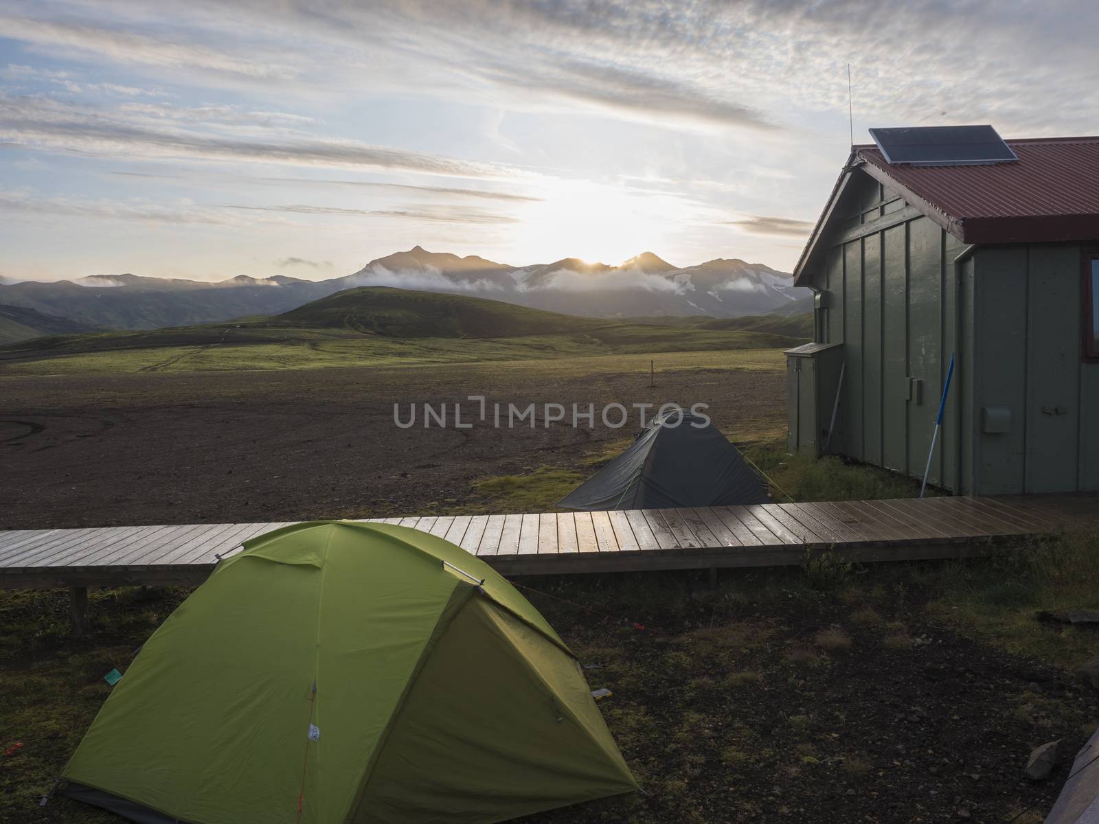 Colorful tents and green tourist hut at Alftavatn camping site with snow covered mountains and green hills and glacier in the background. Landscape of the Fjallabak Nature Reserve in the Highlands of Iceland part of famous Laugavegur hiking trail. Sunrise