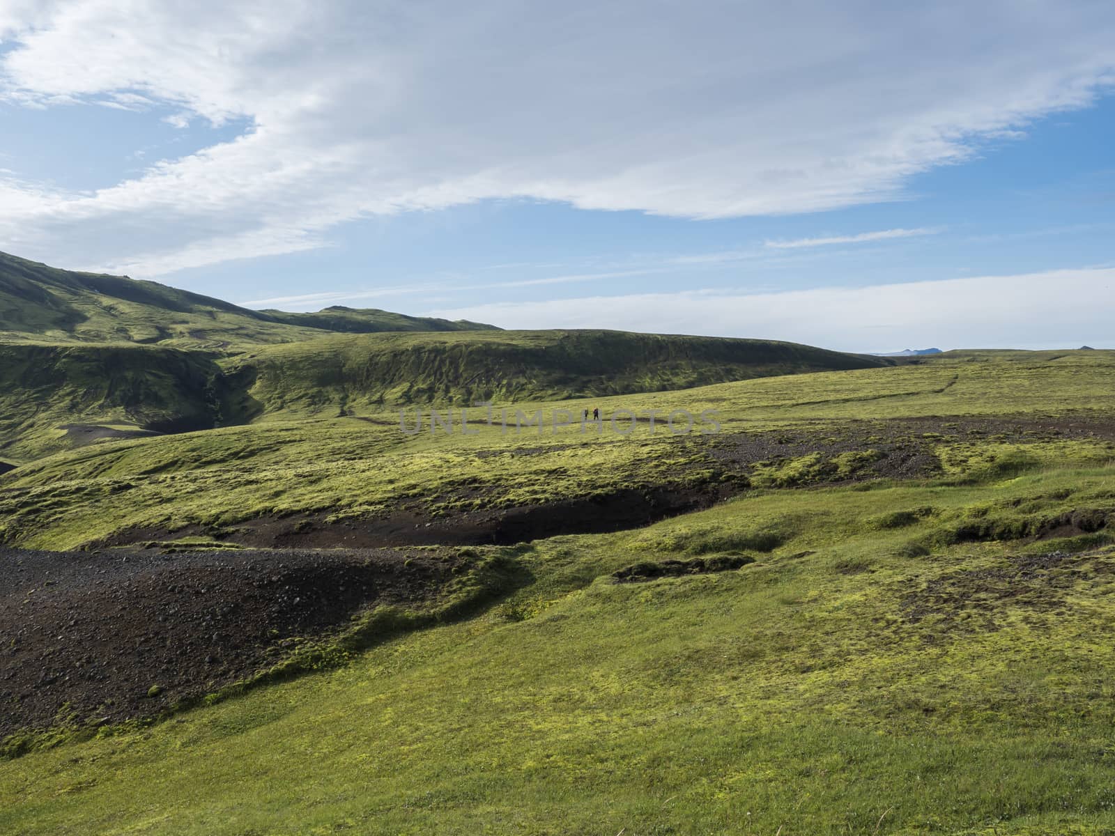 Two hikers backpack travelers in volcanic landscape with green hills, meadow and lava gravel ground covered by lush moss. Fjallabak Nature Reserve in the Highlands of Iceland. Summer blue sky.