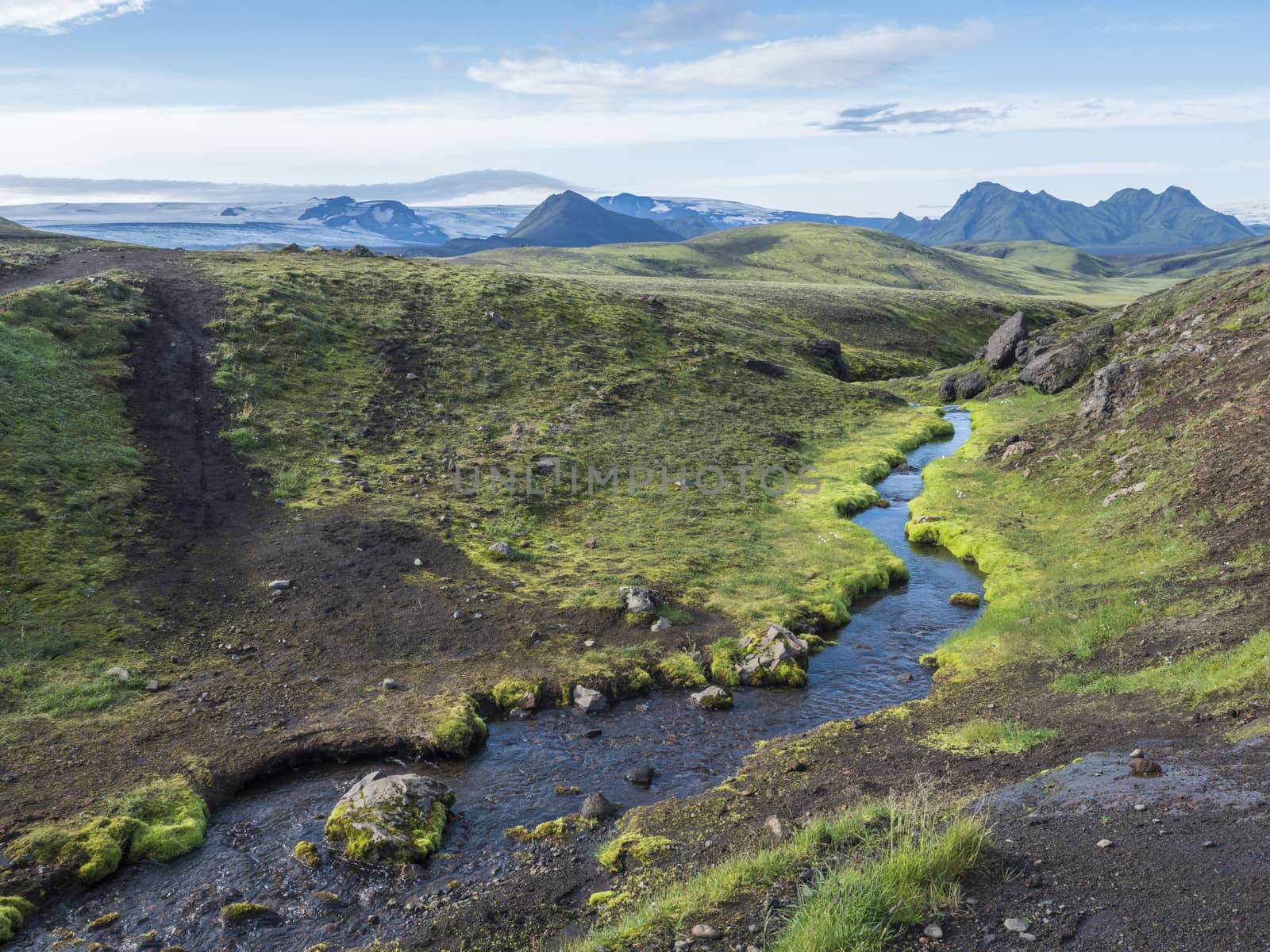 Volcanic landscape with mountains of Tindfjallajokull glacier massif, green hills and blue creek water with lush moss. Fjallabak Nature Reserve, celand. Summer blue sky.