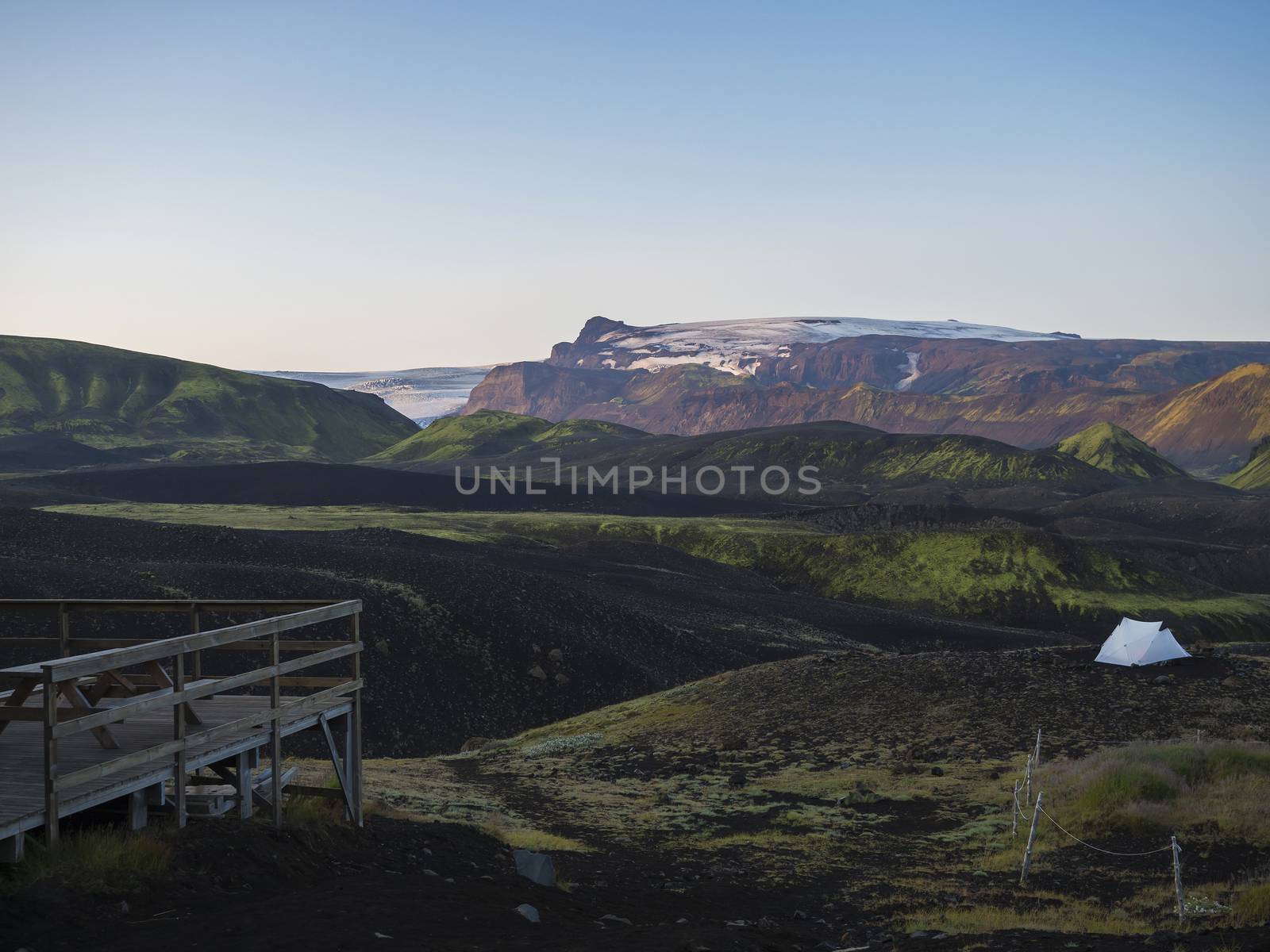Wooden terrace and white tent in Botnar campsite at Iceland on Laugavegur hiking trail, green valley in volcanic landscape with view on Myrdalsjokull glacier. Early morning sunrise pink light.
