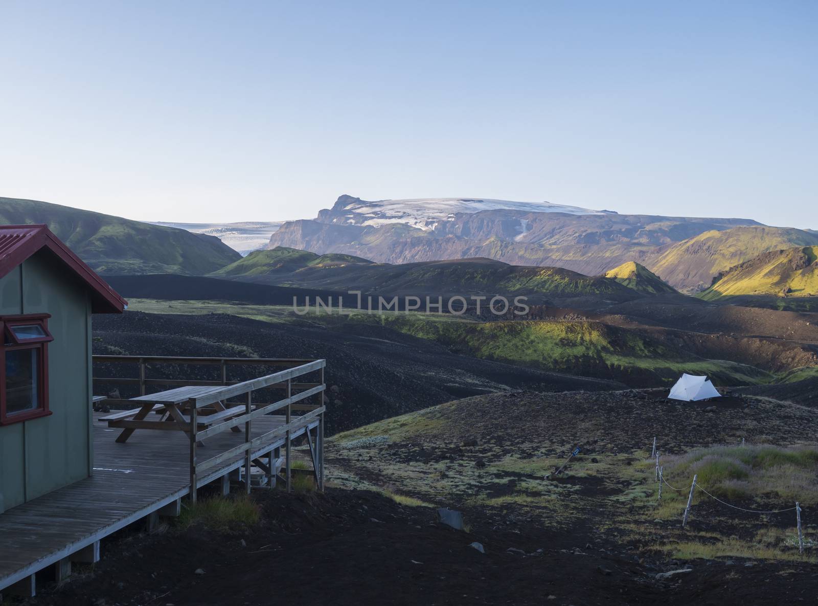 Wooden terrace and white tent in Botnar campsite at Iceland on Laugavegur hiking trail, green valley in volcanic landscape with view on Myrdalsjokull glacier. Early morning sunrise pink light.