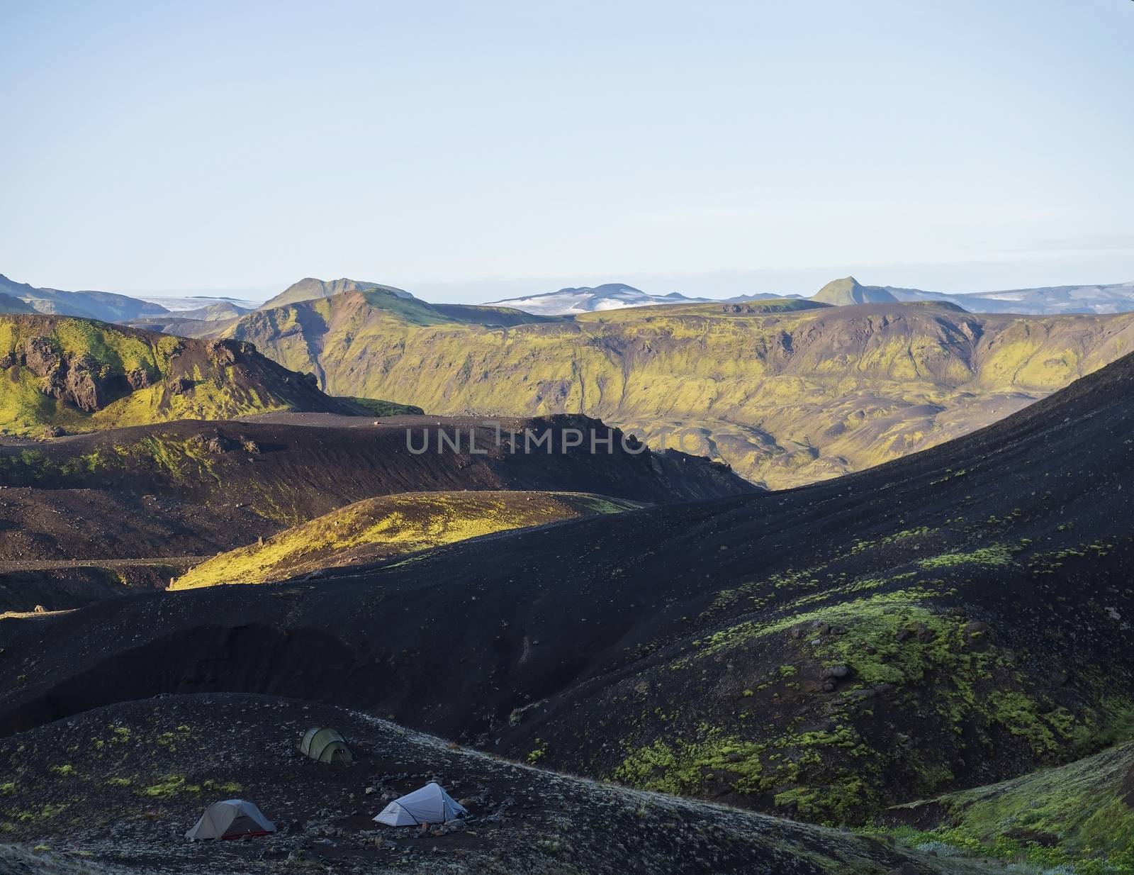 Three tents alone in Botnar campsite at Iceland on Laugavegur hiking trail, green valley in volcanic landscape among lava fields with view on Myrdalsjokull glacier. Early morning sunrise view.