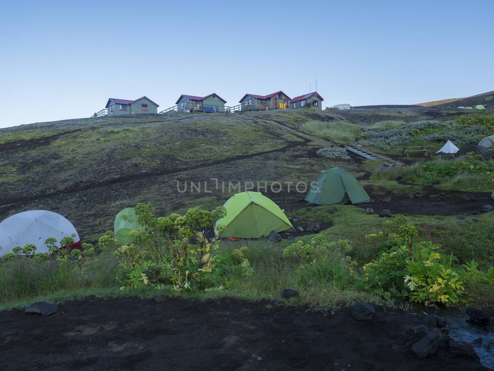 Colorful tents and Botnar mountain hut at Iceland on Laugavegur hiking trail, green valley in volcanic landscape among lava fields with view on Myrdalsjokull glacier. Early morning sunrise view