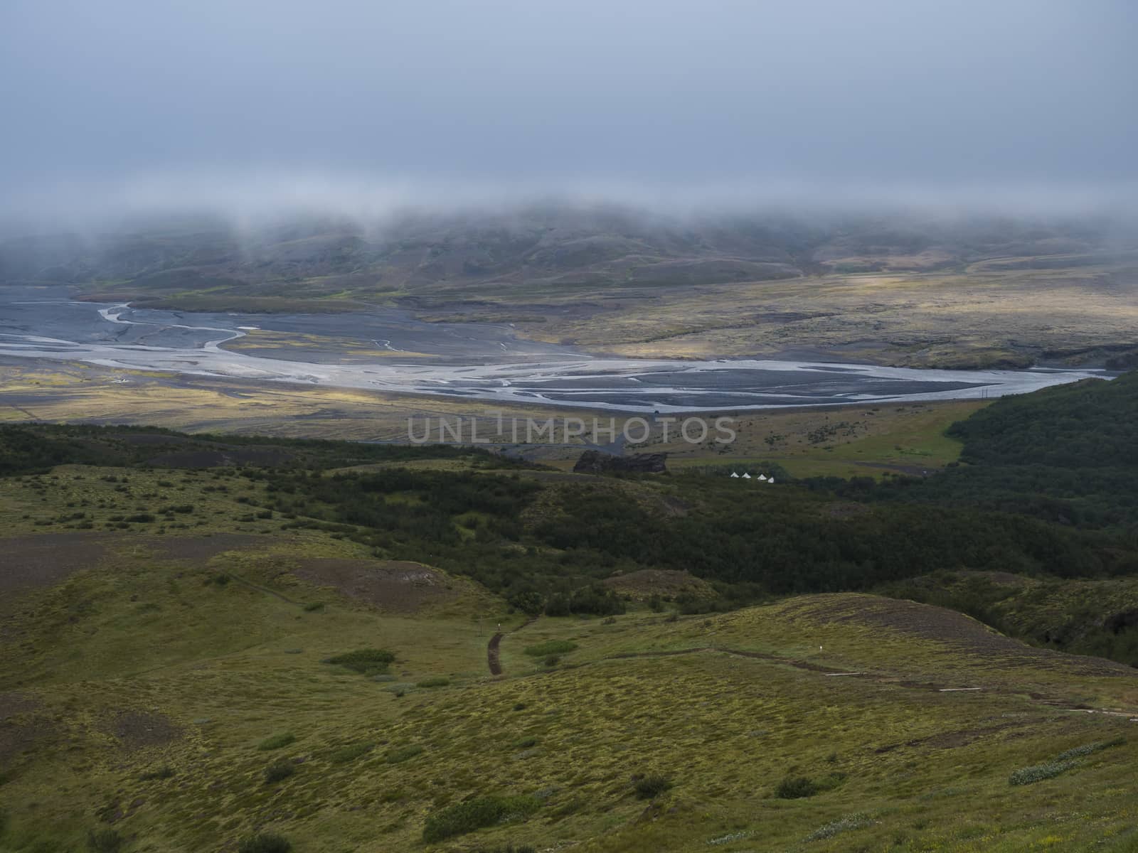 View from top of Valahnukur mountain on landscape of green valley of Thorsmork with volcano huts camp site, sharp green rocks, river Krossa delta and green forest. Foggy summer day, Highlands of Iceland.
