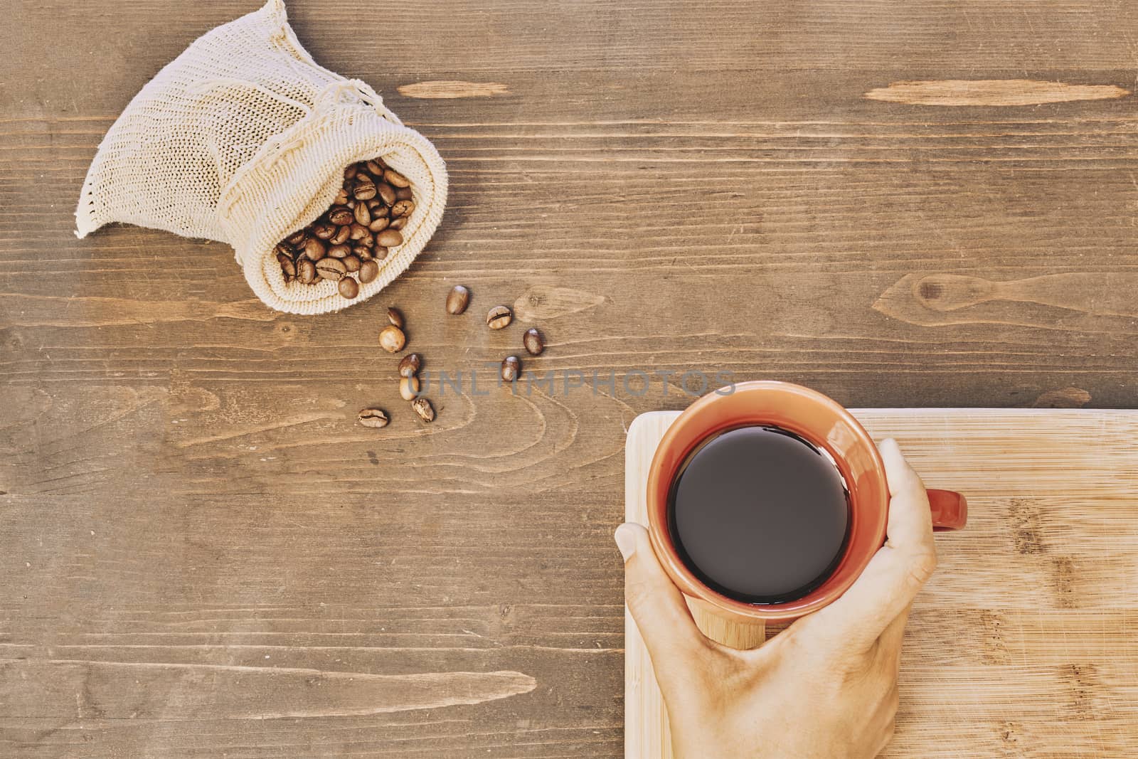 Top view of a person holding a cup of coffee with roasted coffee beans in a small sack on the table