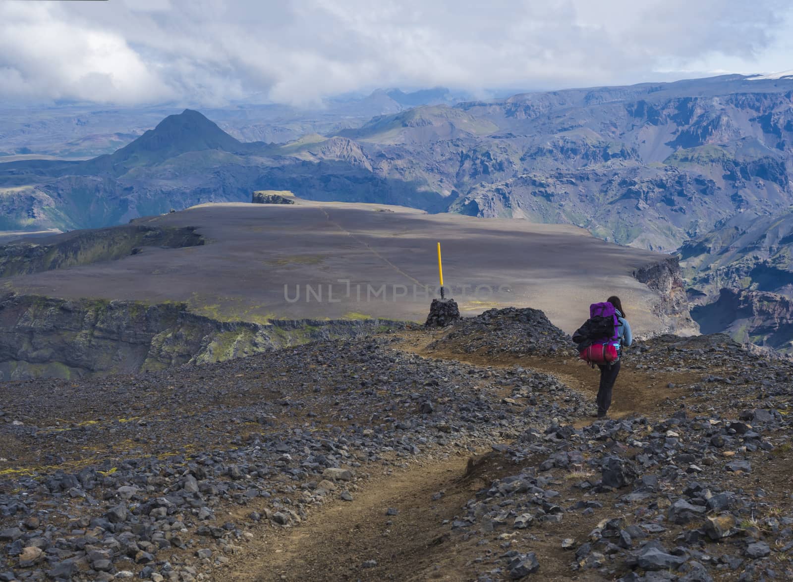 Young woman hiker with backpack at Fimmvorduhals hiking trail. Landscape of Godland with rugged green moss covered rocks and hills, valley of thorsmork, Iceland, Summer blue sky white clouds.