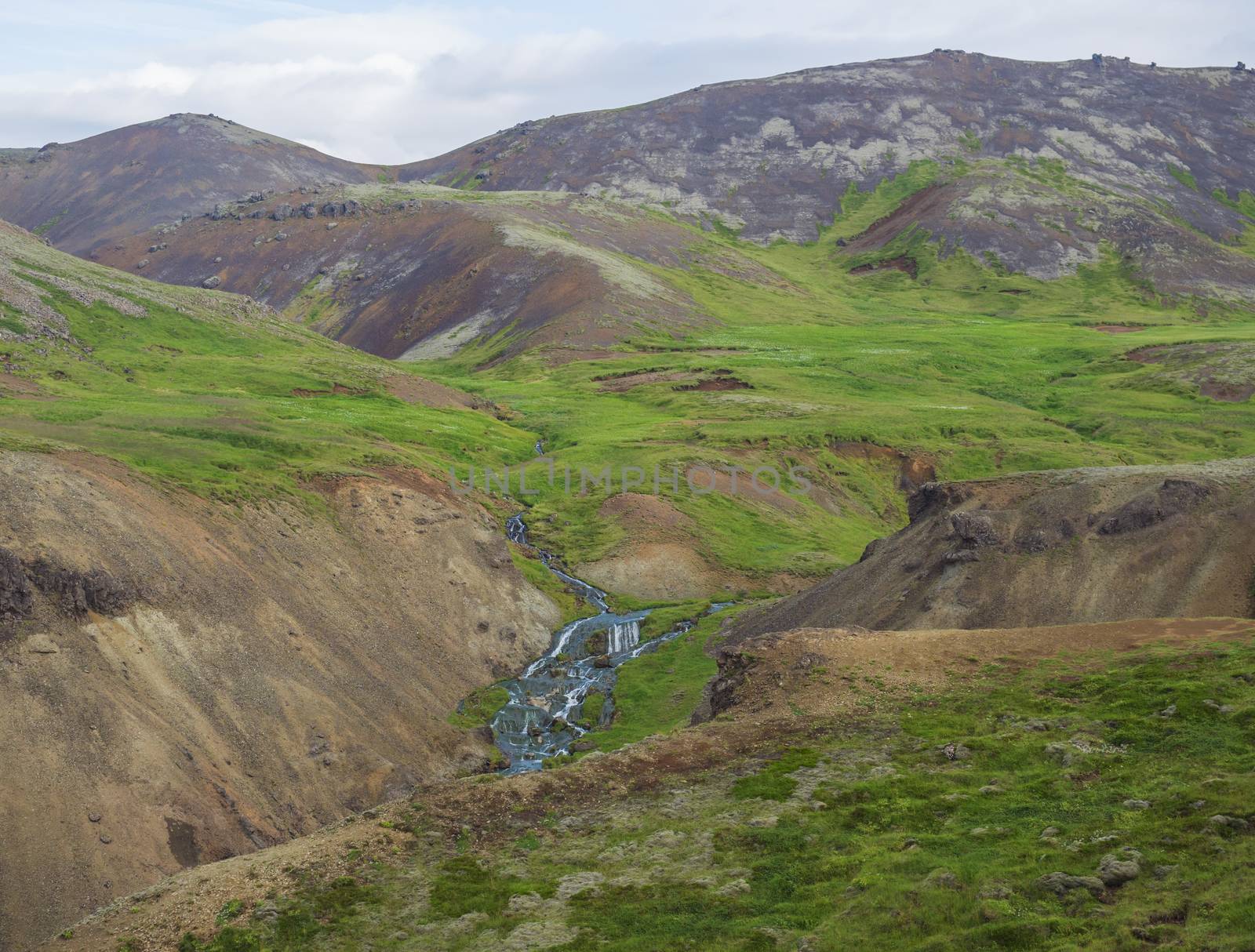 Reykjadalur valley with hot springs river, lush green grass meadow, rocks and hills with geothermal steam. South Iceland near Hveragerdi city. Summer sunny morning, blue sky..