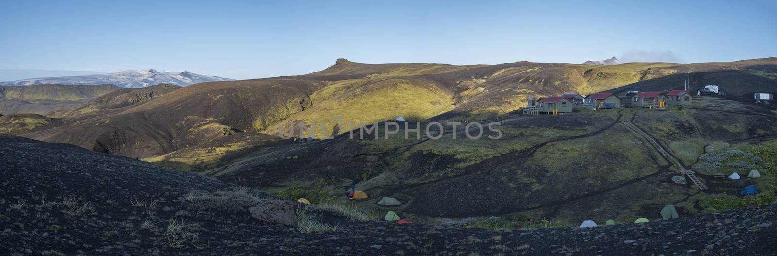 Panoramic view on Botnar mountain hut and campsite at Iceland on Laugavegur hiking trail, green valley in volcanic landscape among lava fields with view on Myrdalsjokull glacier. Early morning, summer blue sky