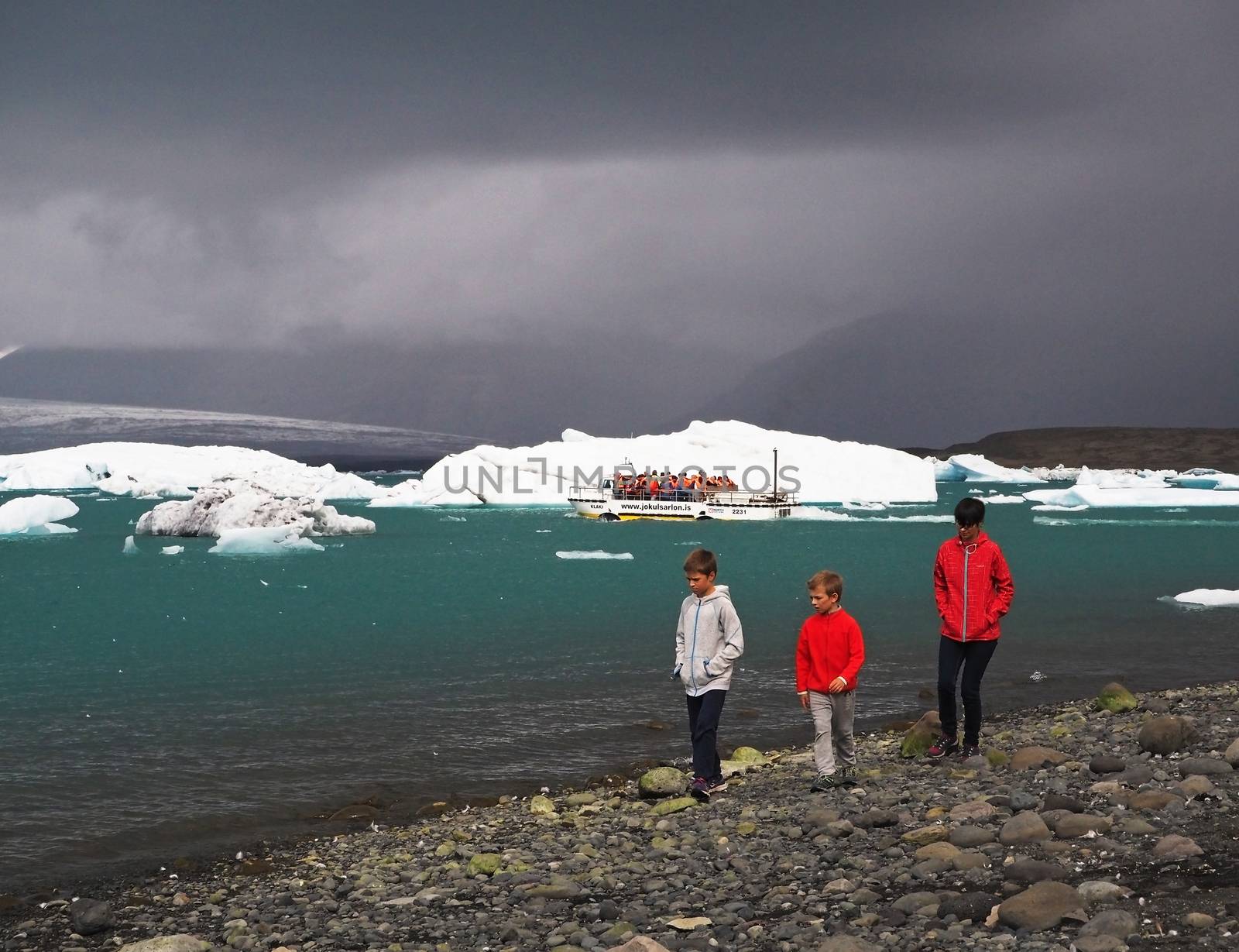 Iceland, Reykjavik, July 3, 2016: two boys and his mather walking in red clothes on the coast line of famous Jokulsarlon lagoon in south eastern Iceland blue white ice blocks and boat with group of tourist in background gray clouds
