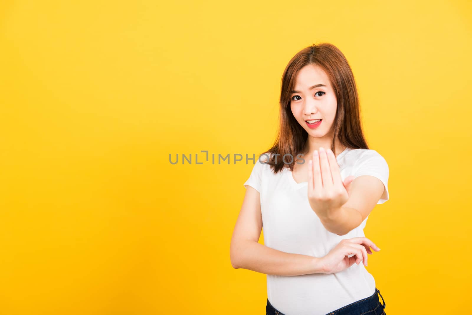 Asian happy portrait beautiful cute young woman teen smile standing wear white t-shirt making gesture hand inviting to come looking to camera isolated, studio shot on yellow background with copy space