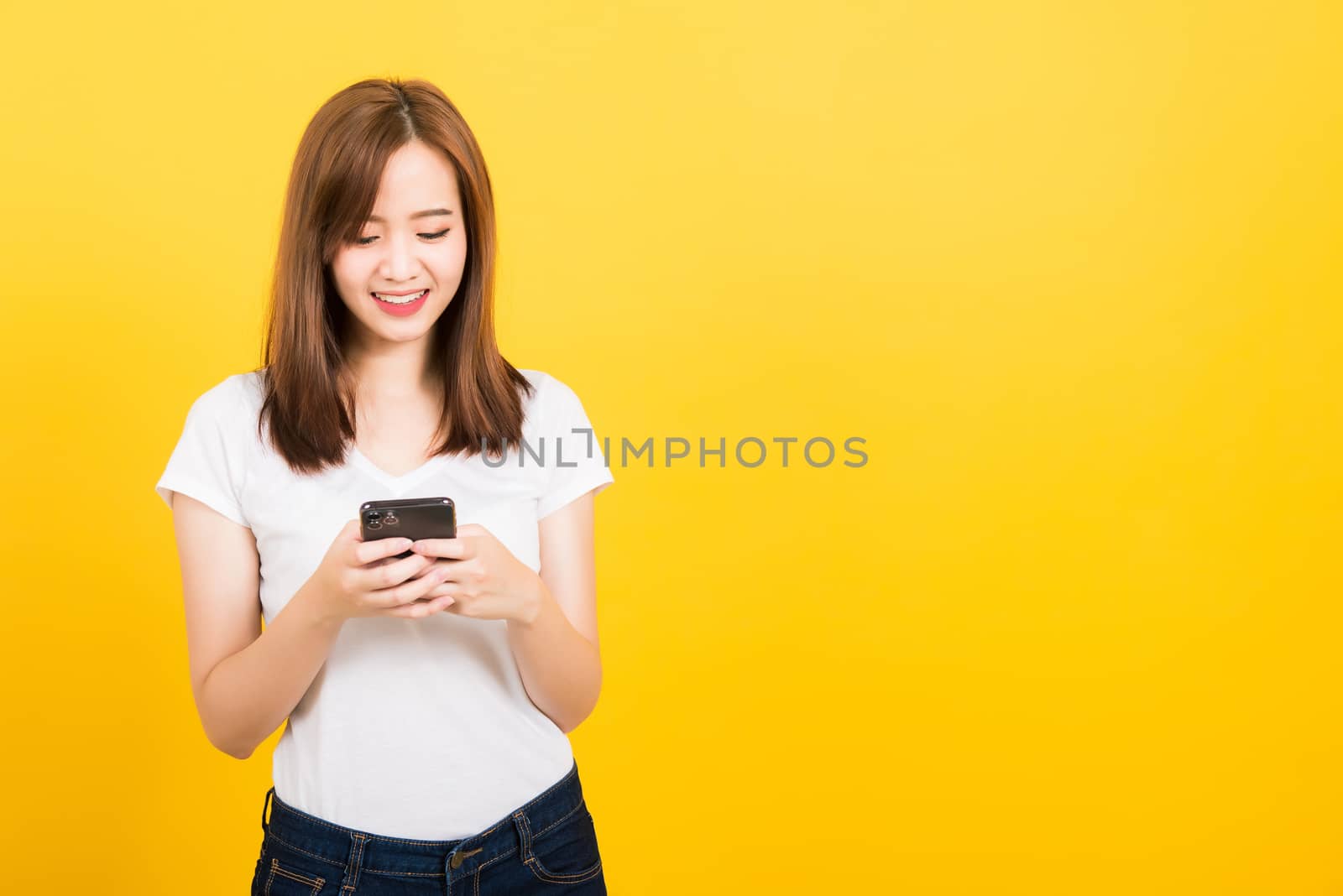 Asian happy portrait beautiful cute young woman teen smile standing playing game or writing message on smartphone looking to the phone isolated, studio shot on yellow background with copy space