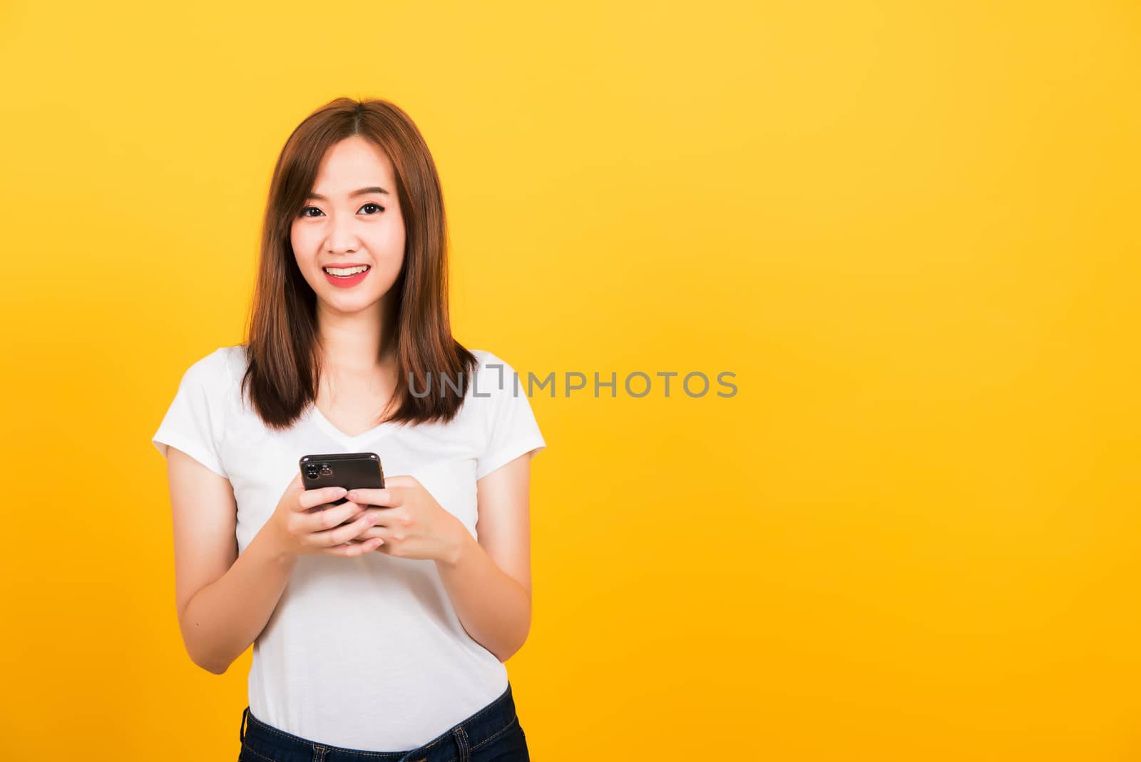 Asian happy portrait beautiful cute young woman teen smile standing playing game or writing message on smartphone looking to camera isolated, studio shot on yellow background with copy space