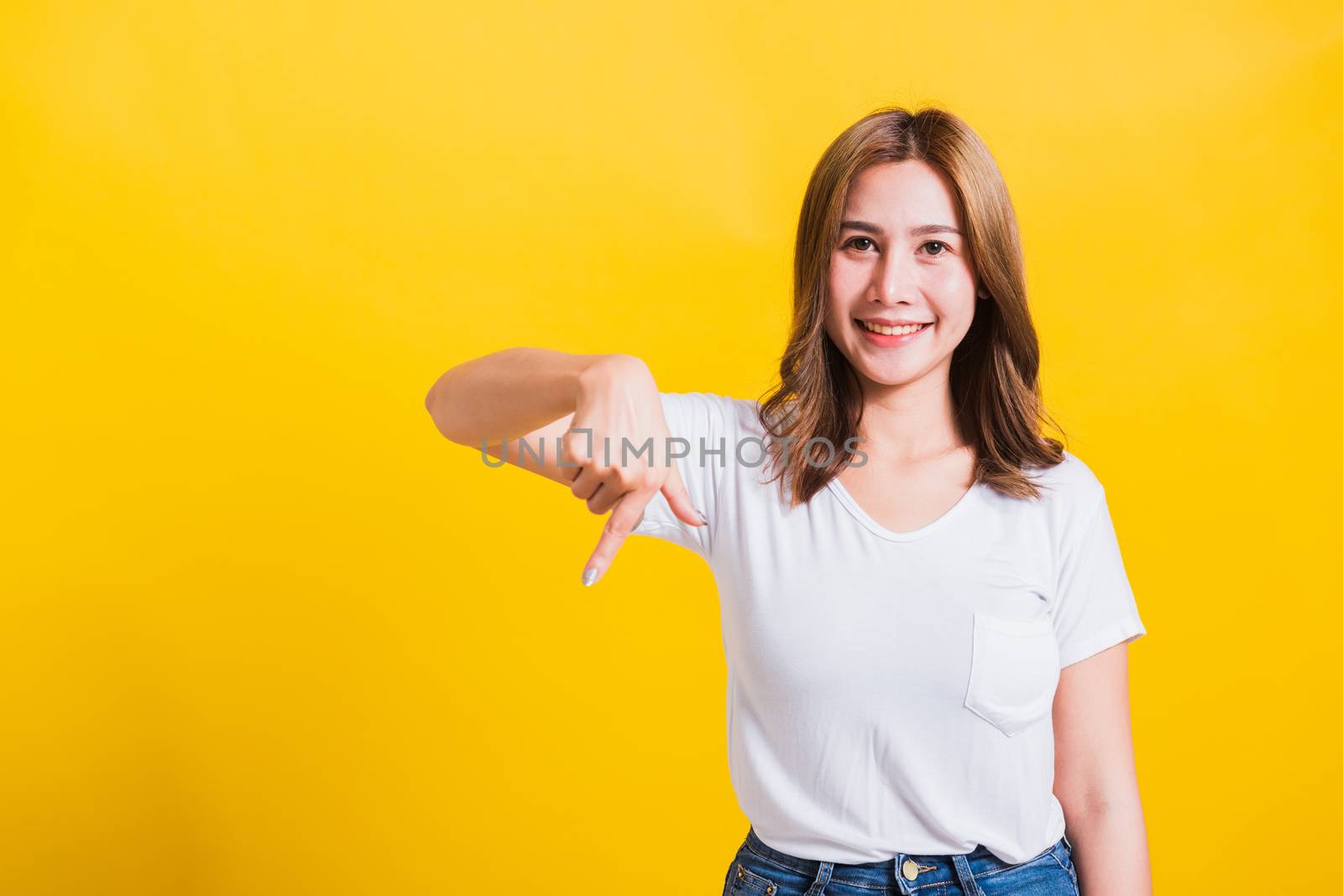 Asian Thai happy portrait beautiful cute young woman standing wear t-shirt makes gesture two fingers point below down and looking to camera, studio shot isolated on yellow background with copy space