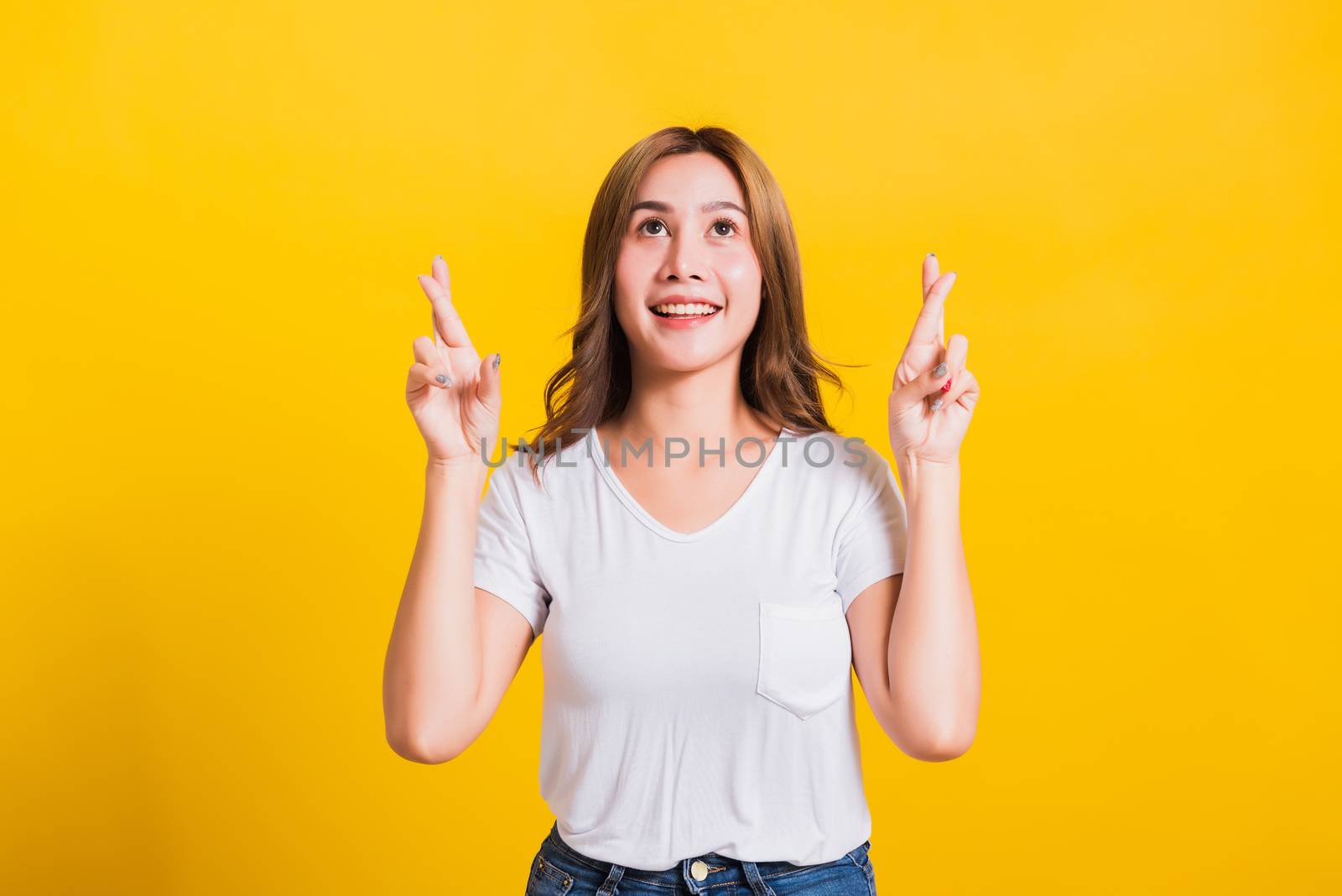 Asian Thai happy portrait beautiful cute young woman smile have superstition her holding fingers crossed for good luck and looking to camera, studio shot isolated on yellow background with copy space