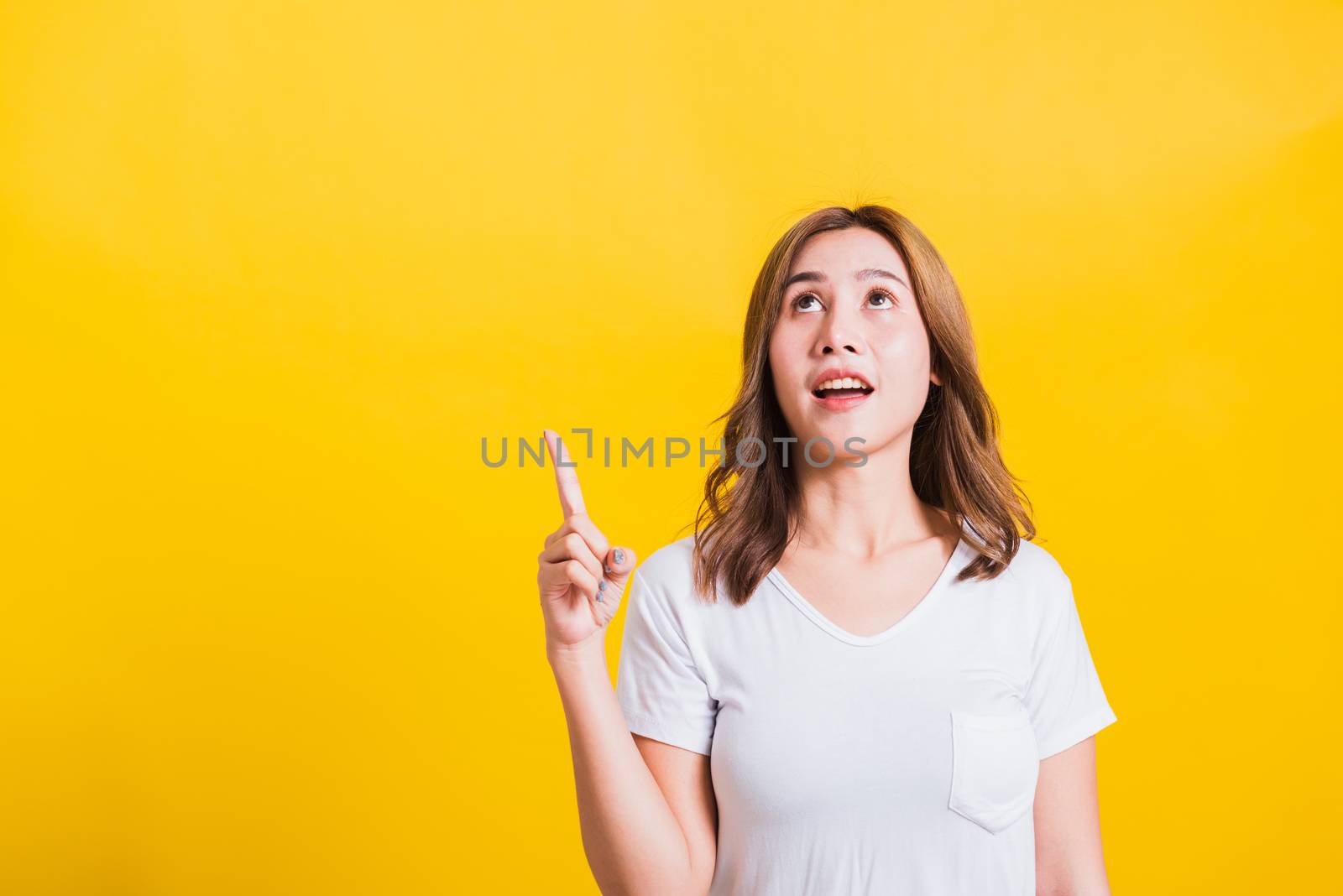 Asian Thai happy portrait beautiful cute young woman standing wear white t-shirt makes gesture two fingers point upwards above looking above, studio shot isolated on yellow background with copy space