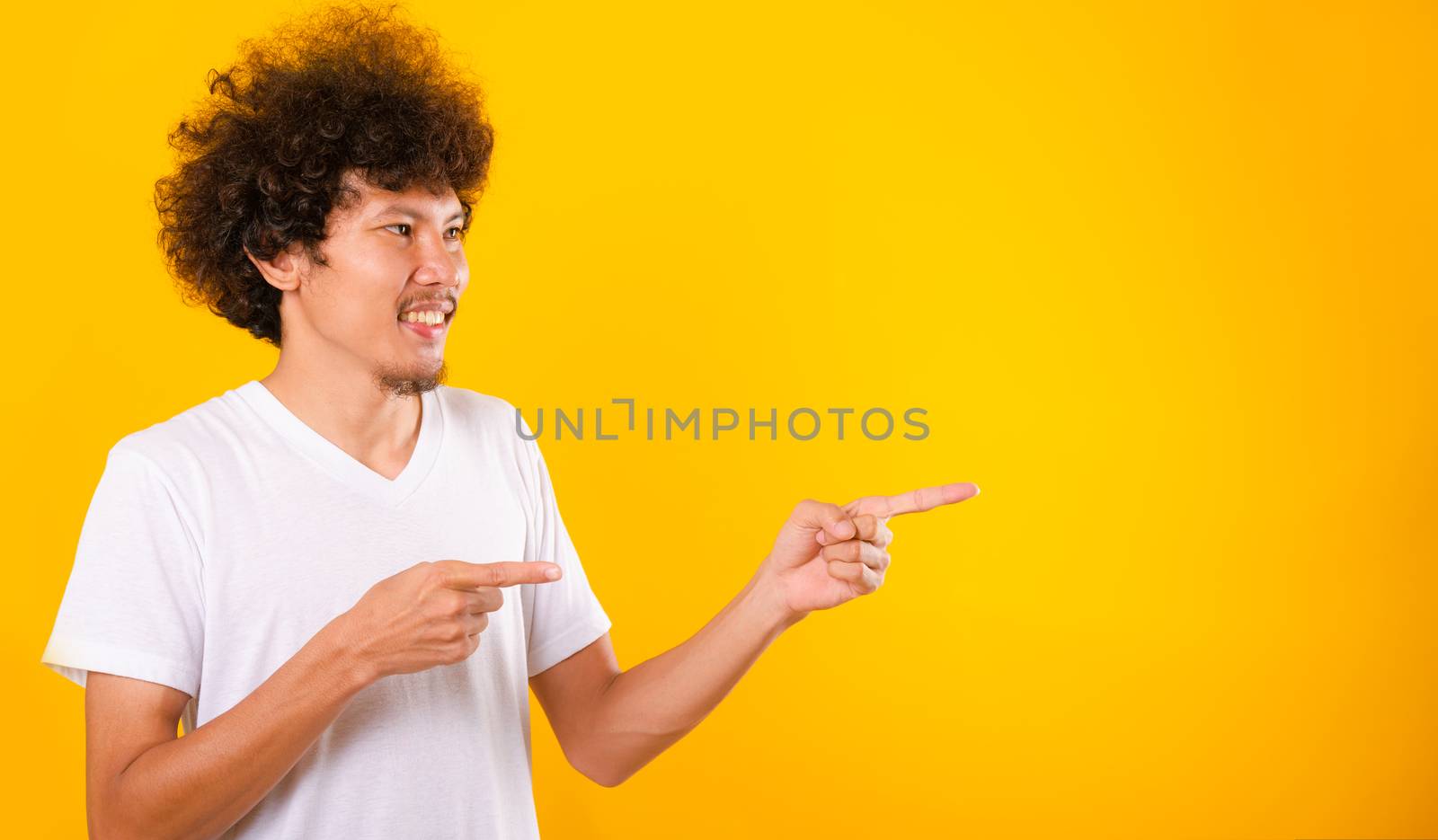 Asian Portrait happy young man curly hair White t-shirt pointing fingers away copy space isolated yellow background