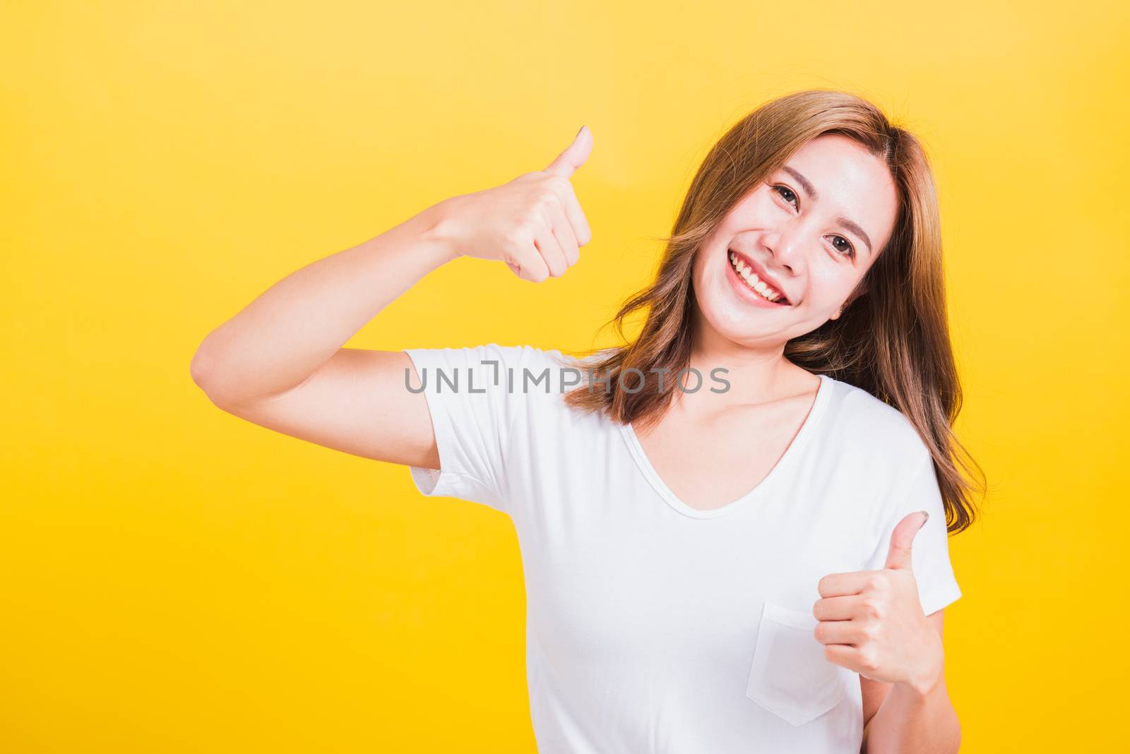 Portrait Asian Thai beautiful happy young woman smiling wear white t-shirt standing successful woman giving two thumbs up gesture sign in studio shot, isolated on yellow background with copy space