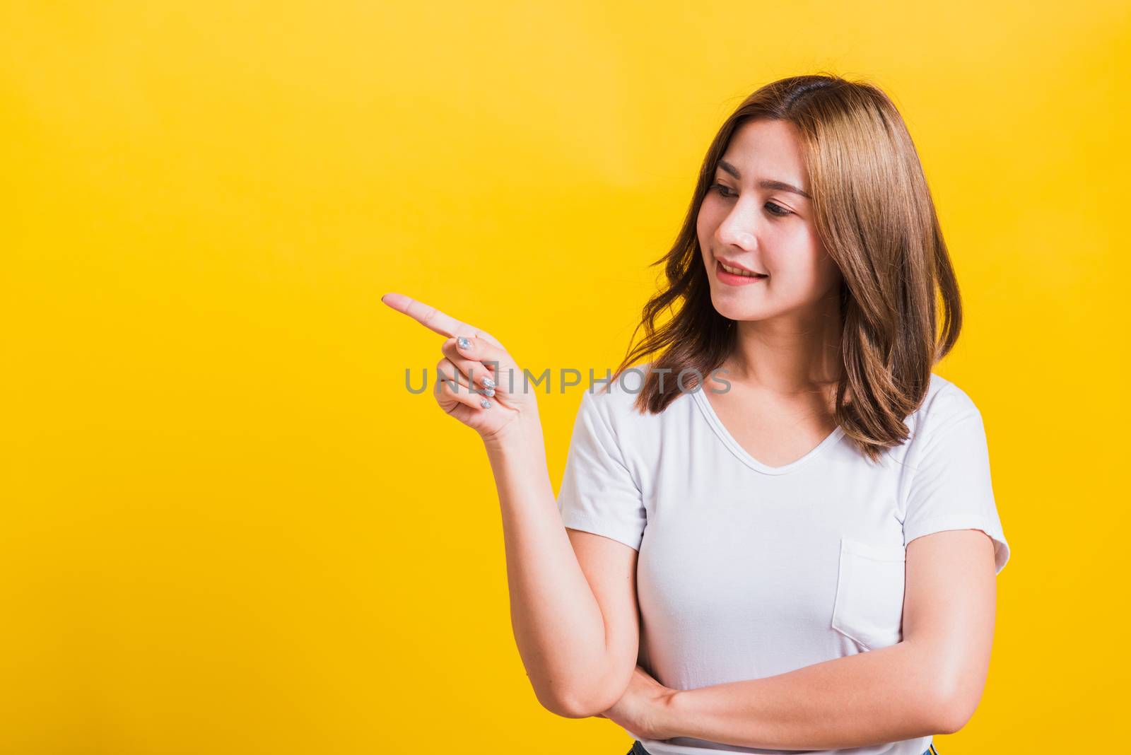 Asian Thai happy portrait beautiful cute young woman standing wear white t-shirt pointing finger away side looking to away side, studio shot isolated on yellow background with copy space