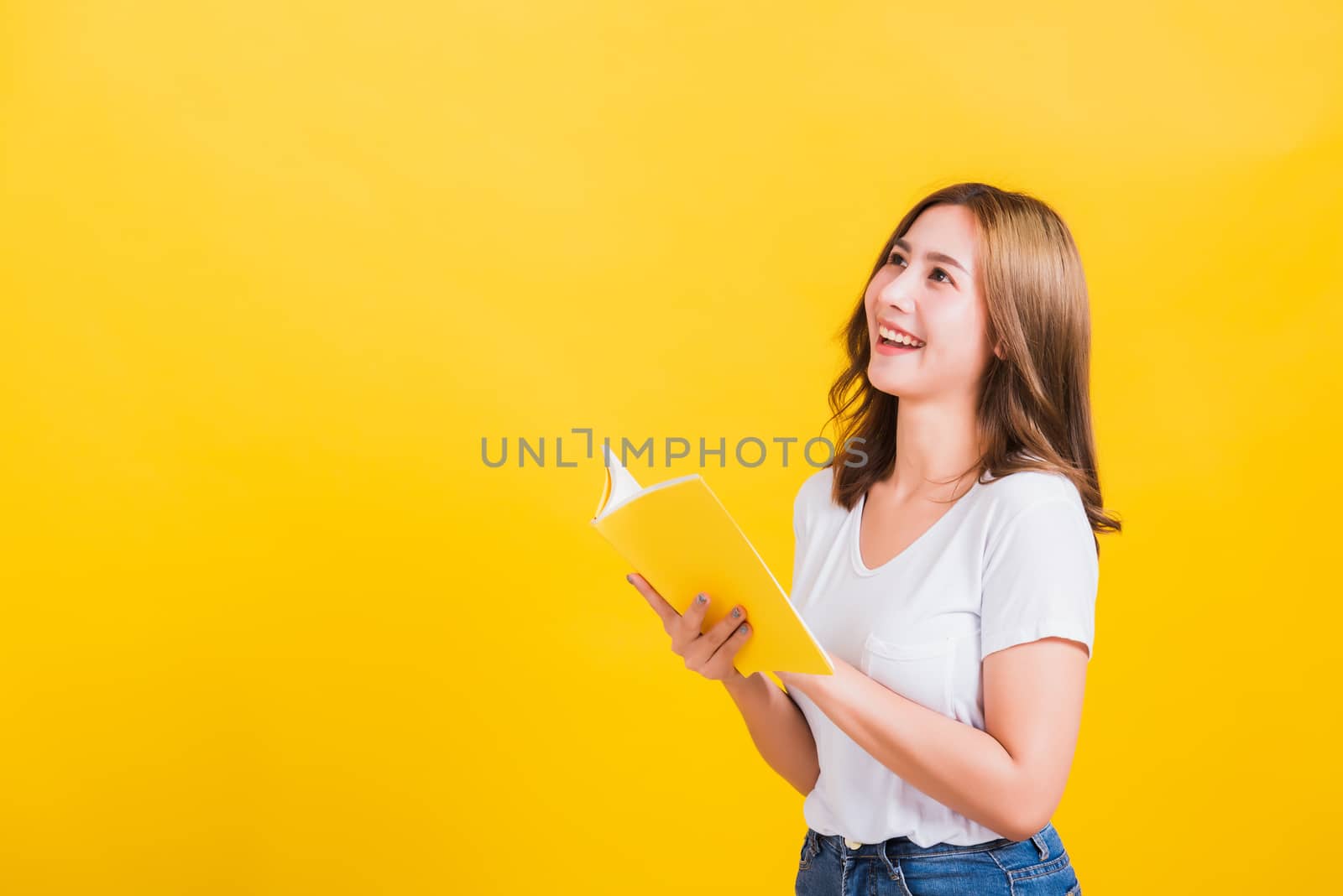 Portrait Asian Thai beautiful happy young lifestyle woman stands holding yellow book or diary she show thumb up finger and looking up away, studio shot isolated on yellow background, with copy space
