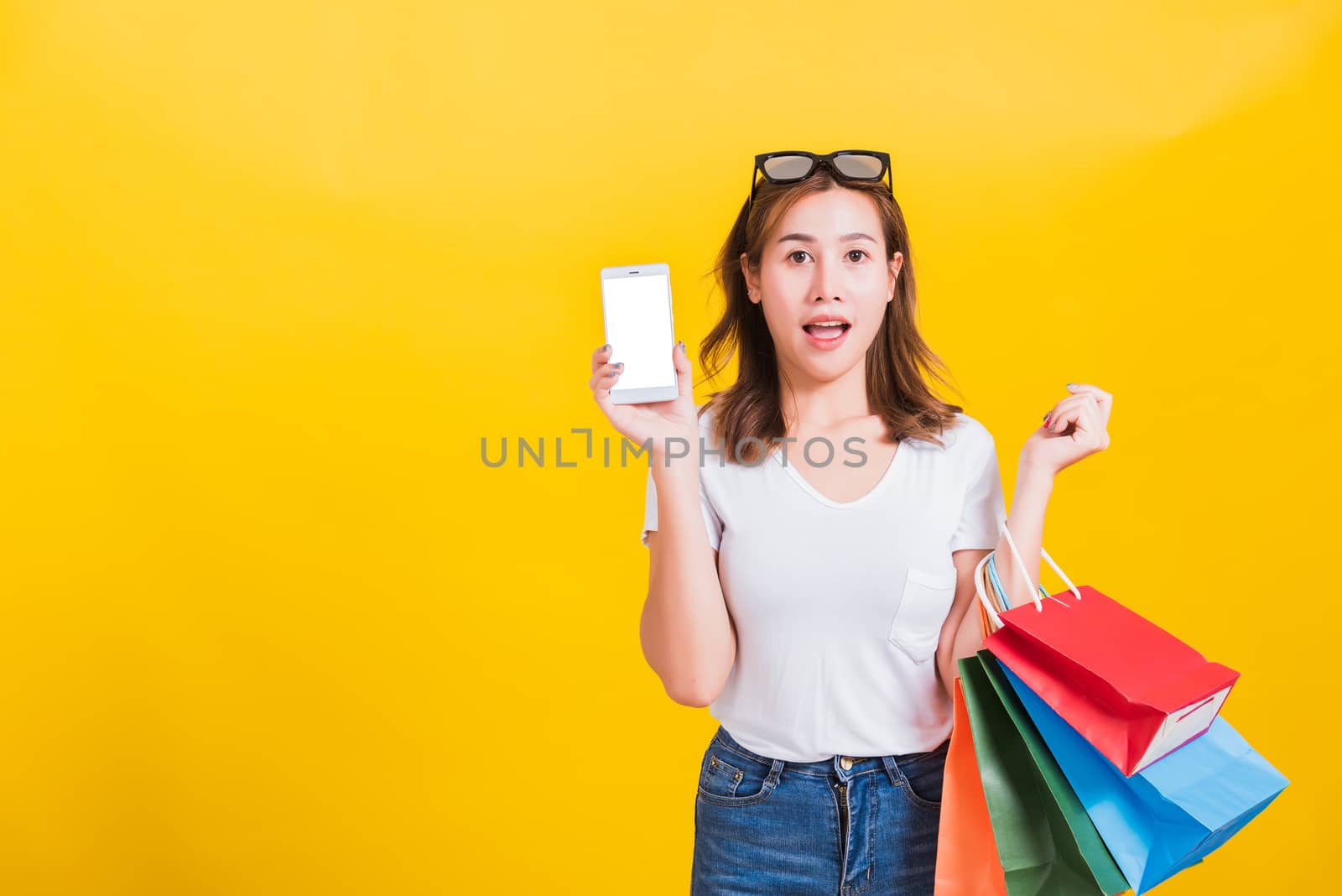 Portrait happy Thai Asian beautiful young woman smile standing, she surprises open mouth and holding shopping bags and show mobile phone blank screen, studio shot on yellow background with copy space
