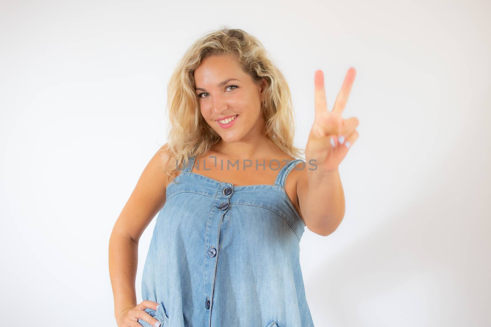 Pretty blonde woman in blue dress smiling making victory gesture