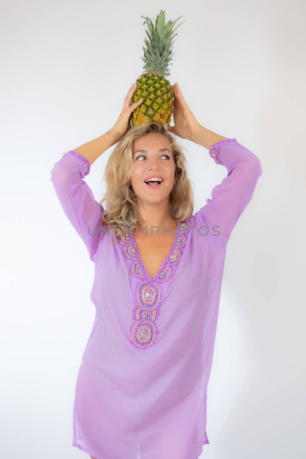 Pretty blonde woman in lila caftan smiling on white background picking up a pineapple on her head