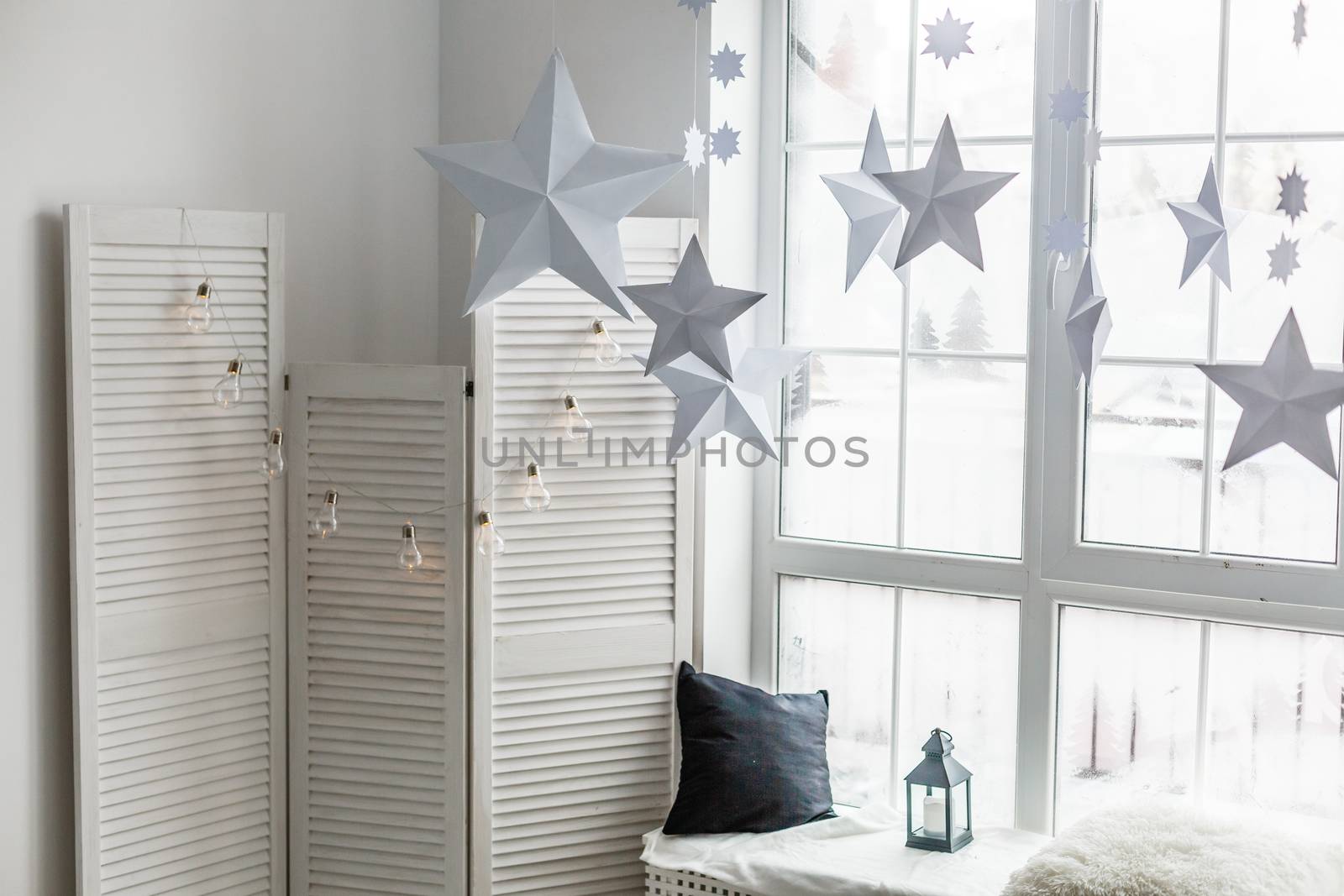 Large bright window. On the window-sill is a gray knitted plaid with gifts and sparkles with herlands. Paper Christmas stars are suspended on the background of the window. by Andelov13