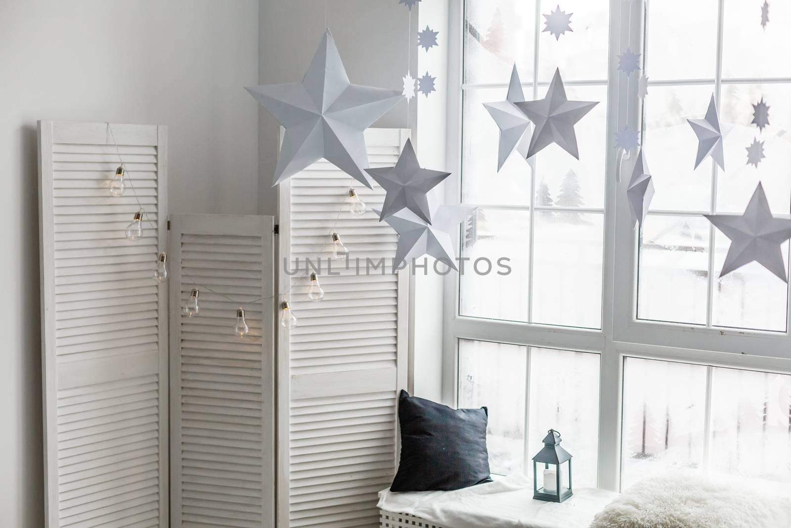 Large bright window. On the window-sill is a gray knitted plaid with gifts and sparkles with herlands. Paper Christmas stars are suspended on the background of the window.