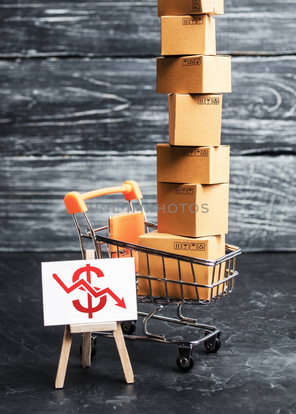 A shopping trolley loaded with boxes and an easel with a red dollar down arrow. Internet trade, online purchase. Drop in sales, economic recession. Retail of goods and services, search for new markets