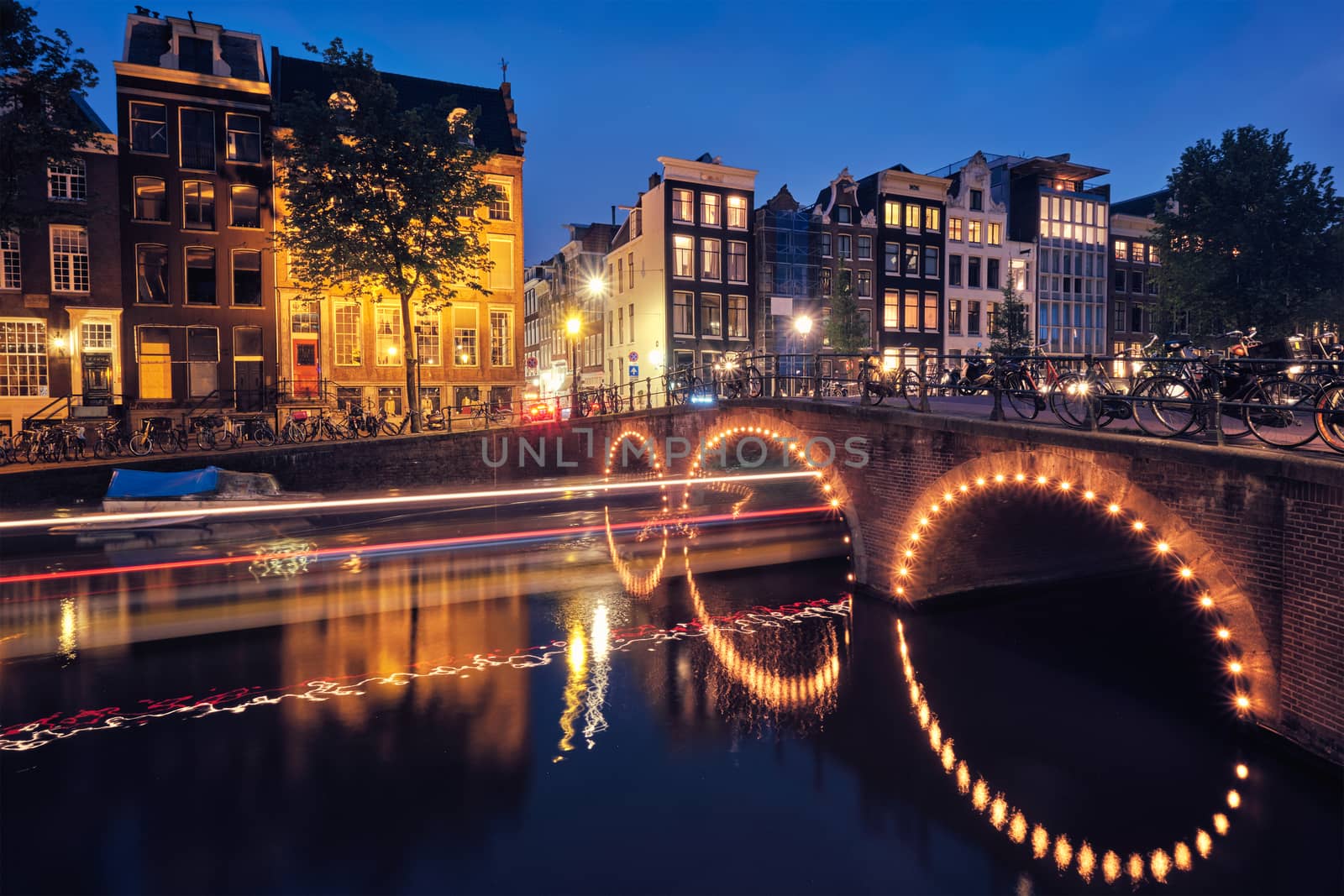 Night view of Amterdam cityscape with canal, bridge and medieval houses in the evening twilight illuminated with boat light trails. Amsterdam, Netherlands