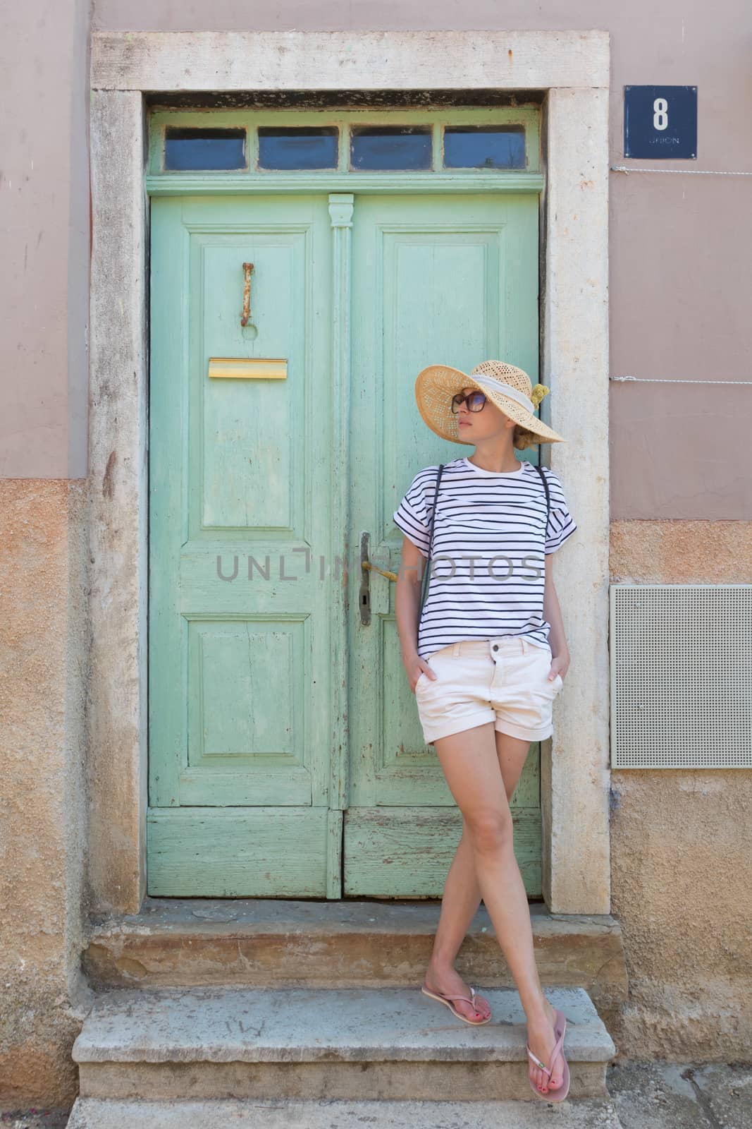 Beautiful young female tourist woman wearing sun hat, standing and relaxing in shade in front of turquoise vinatage wooden door in old Mediterranean town while sightseeing on hot summer day.