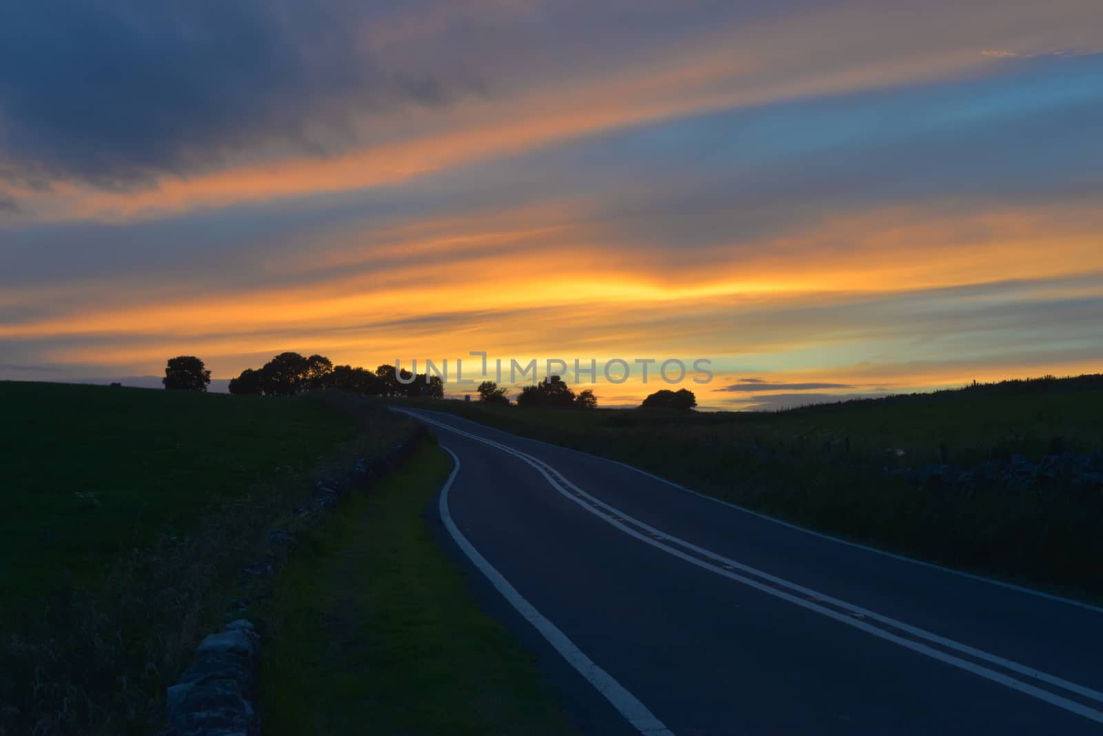 Deep Blue fresh tarmac road in the peak district leads to some trees and a strong orange sunset.